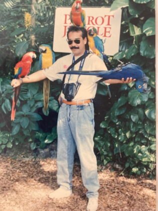 Man with macaws perched on his arms in front of a Parrot Jungle sign.