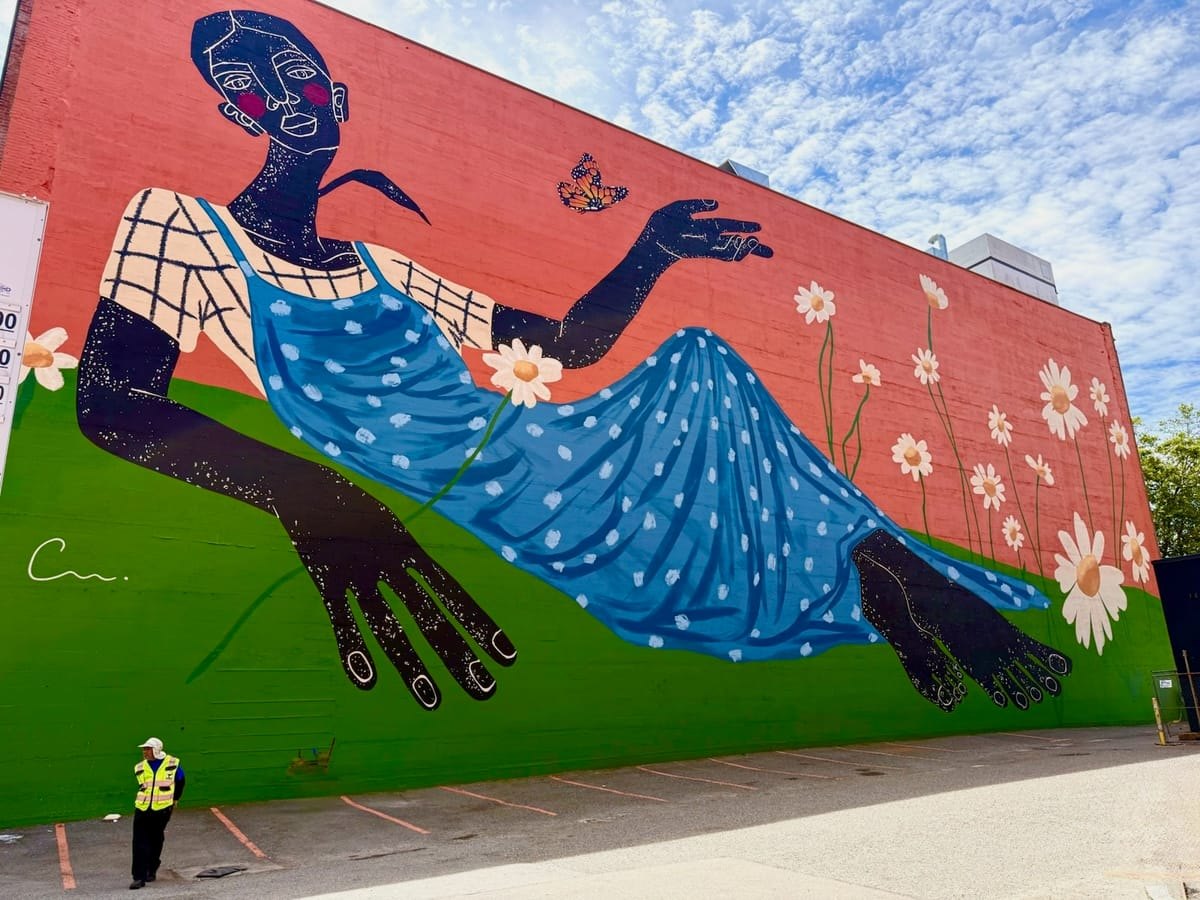 Large outdoor mural on a brick building showing a reclining woman in a blue dress surrounded by daisies.