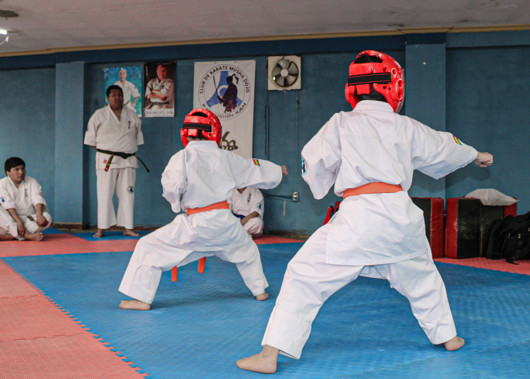 Two children in white karate uniforms and red headgear sparring on a tatami mat.
