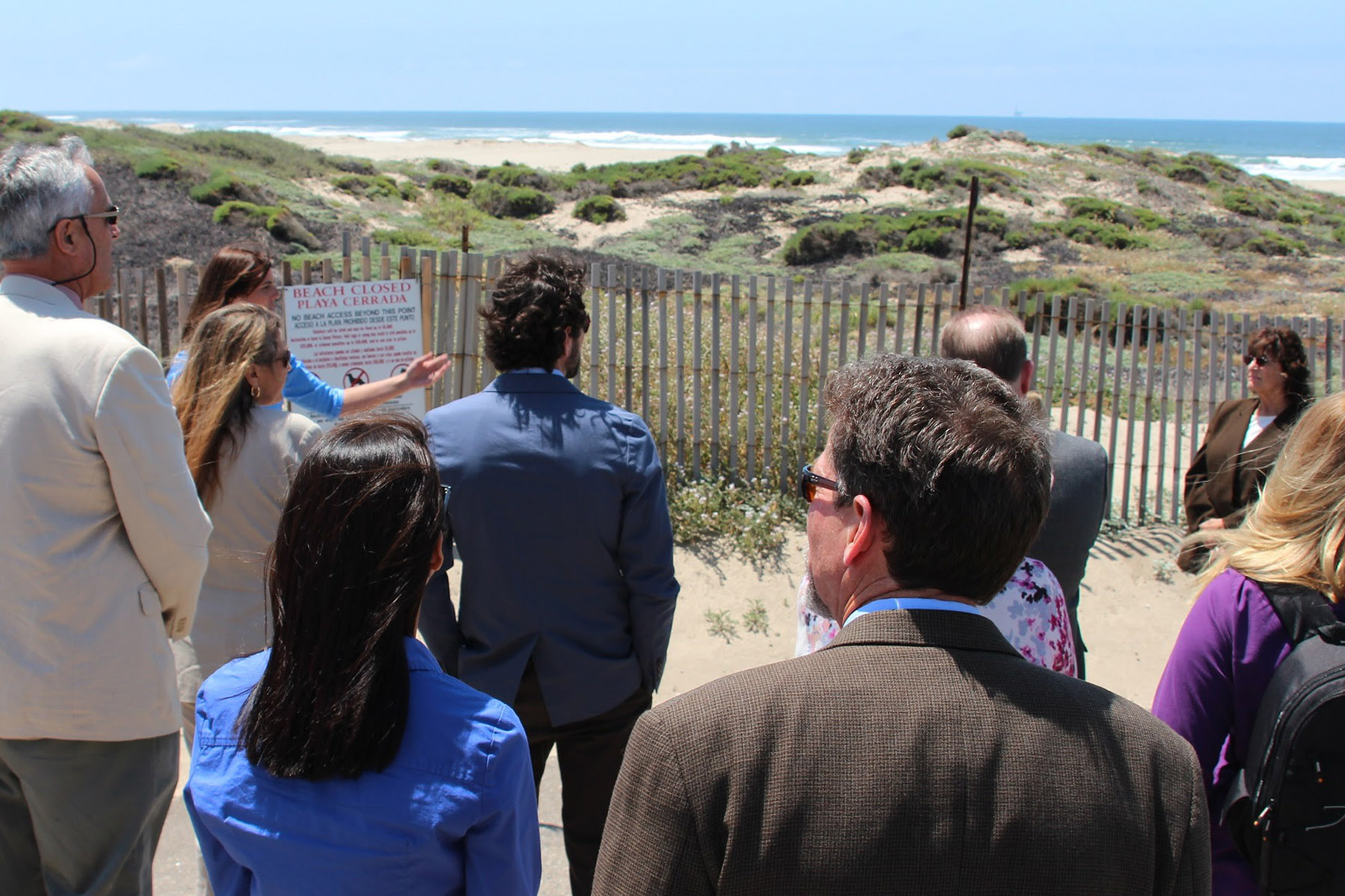 Officials in business attire gathered near a coastal fence with ocean and dunes visible.