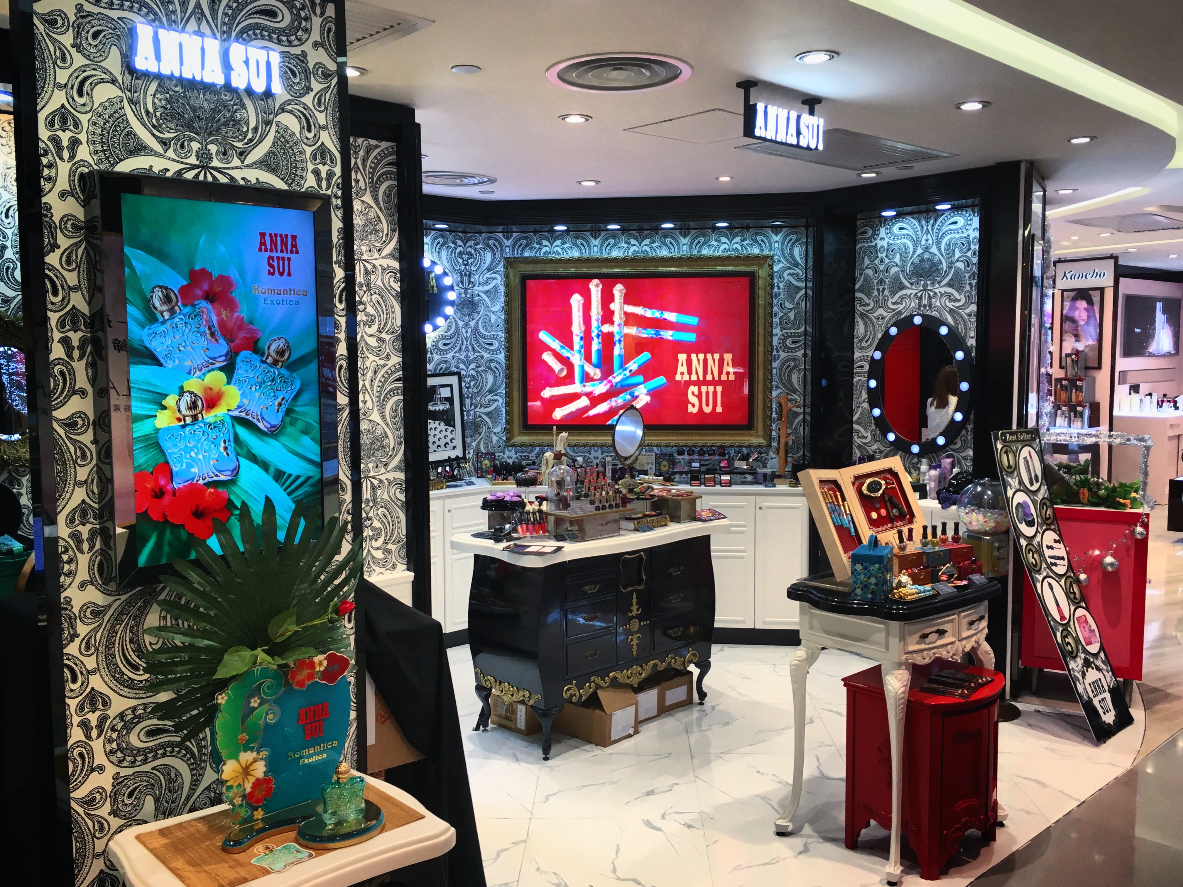 Anna Sui store interior with black damask wallpaper, illuminated logo signage, and cosmetics display cases.