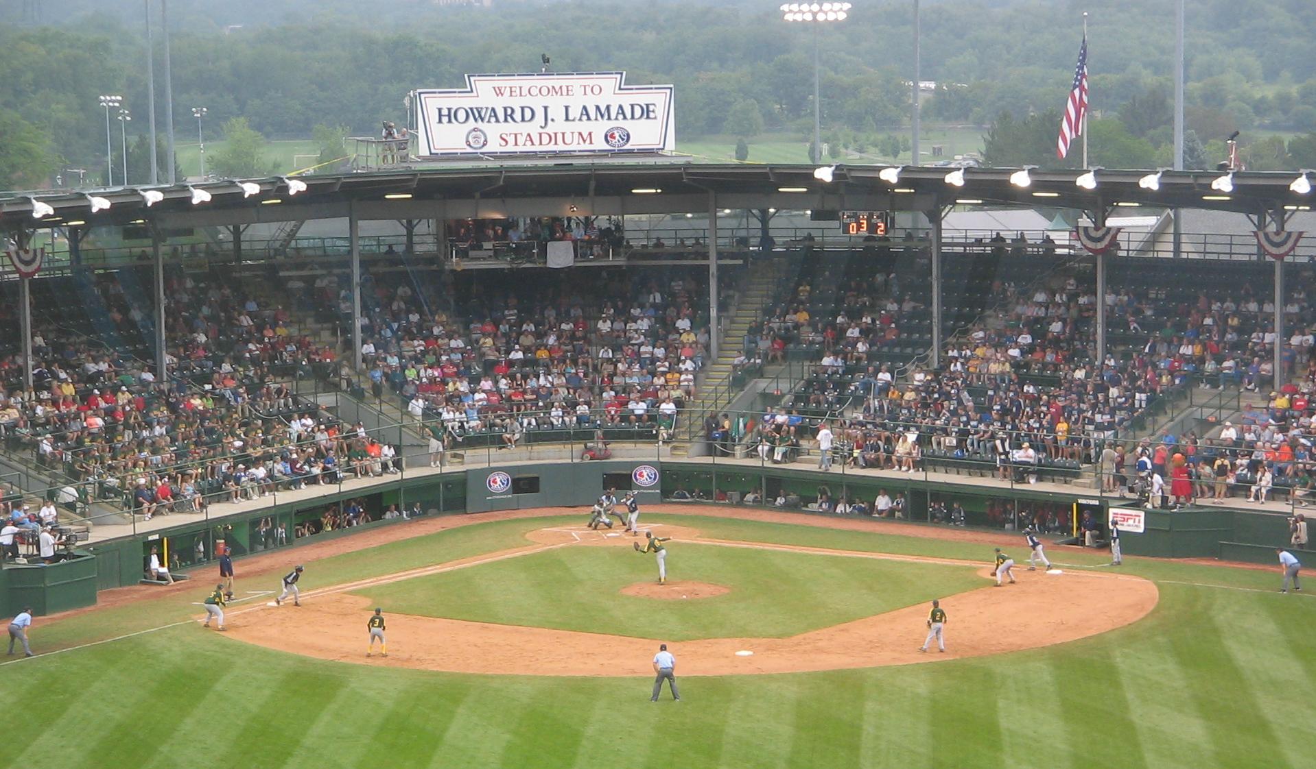 Youth baseball game at a packed Little League stadium with an American flag flying above.