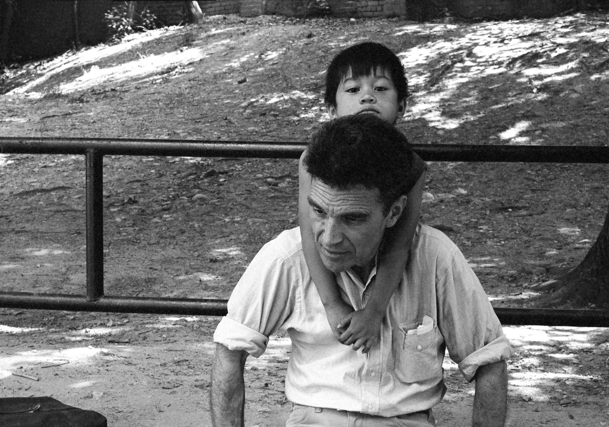 An older man in a light shirt with a young child's arms draped over his shoulders from behind, both appearing tired, outdoors.