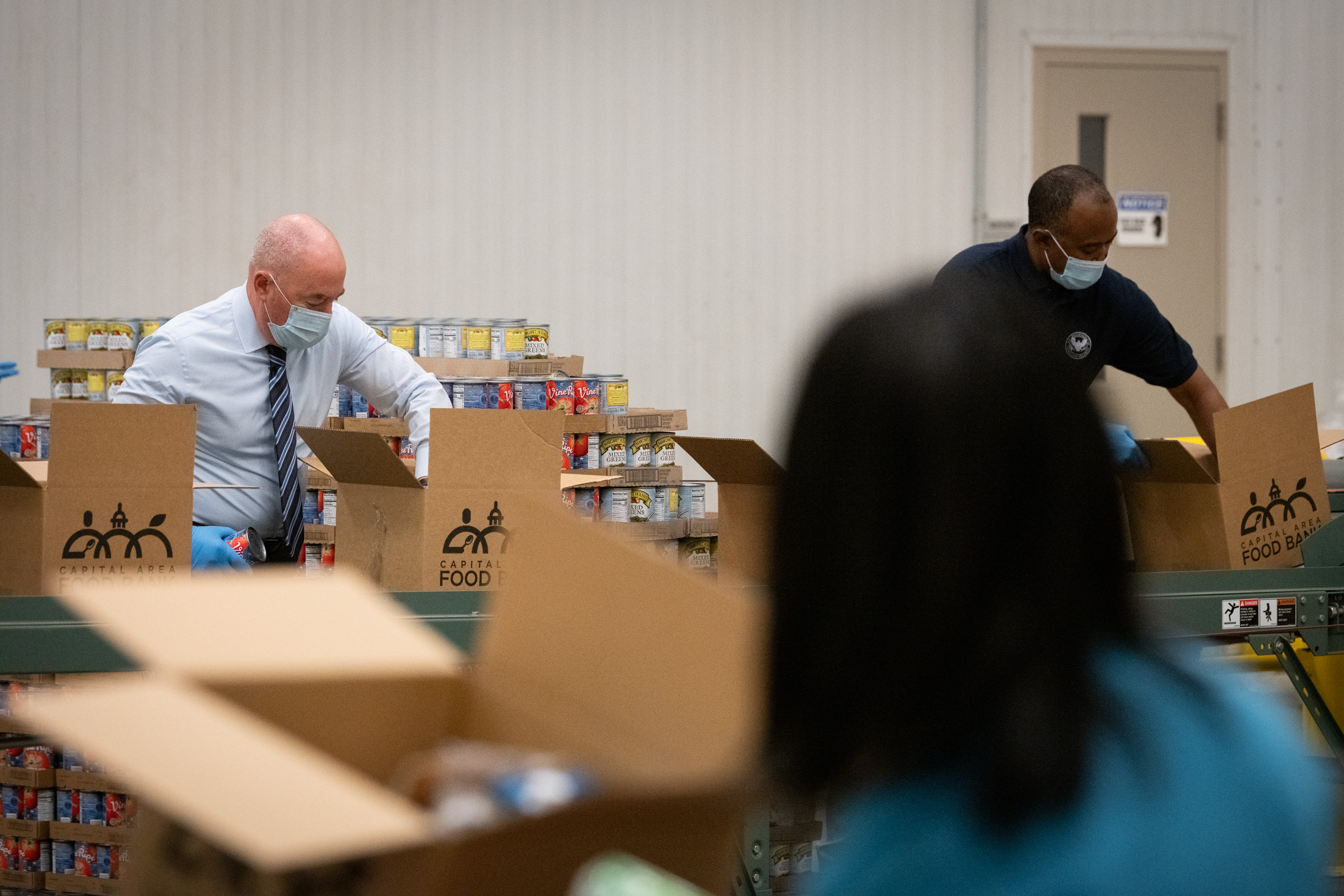 Two masked volunteers in gloves pack canned goods into labeled food bank boxes.