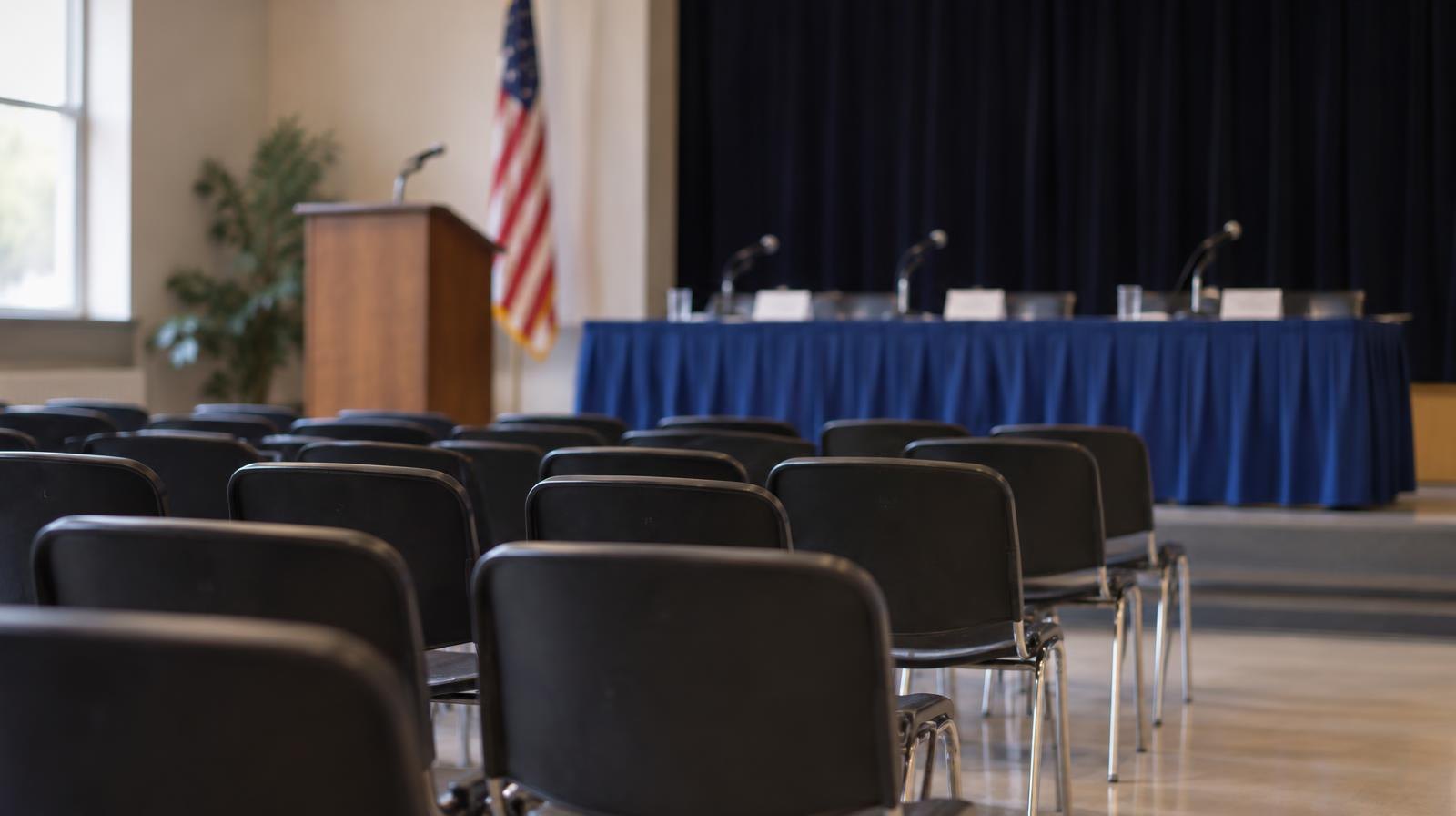 Empty room with rows of chairs facing a panel table at the front.