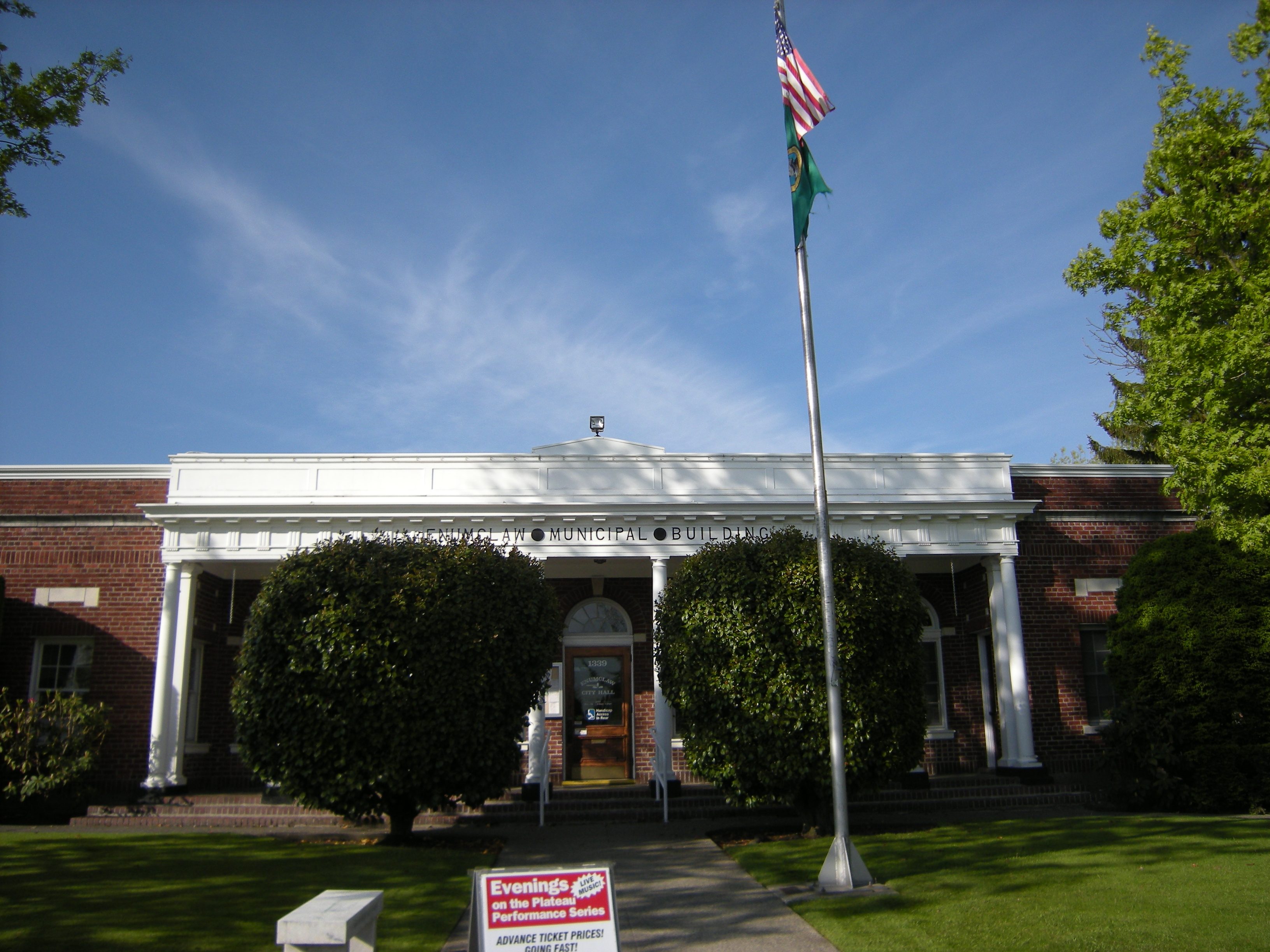 Brick city hall building with white columned entrance and two flags on a flagpole.
