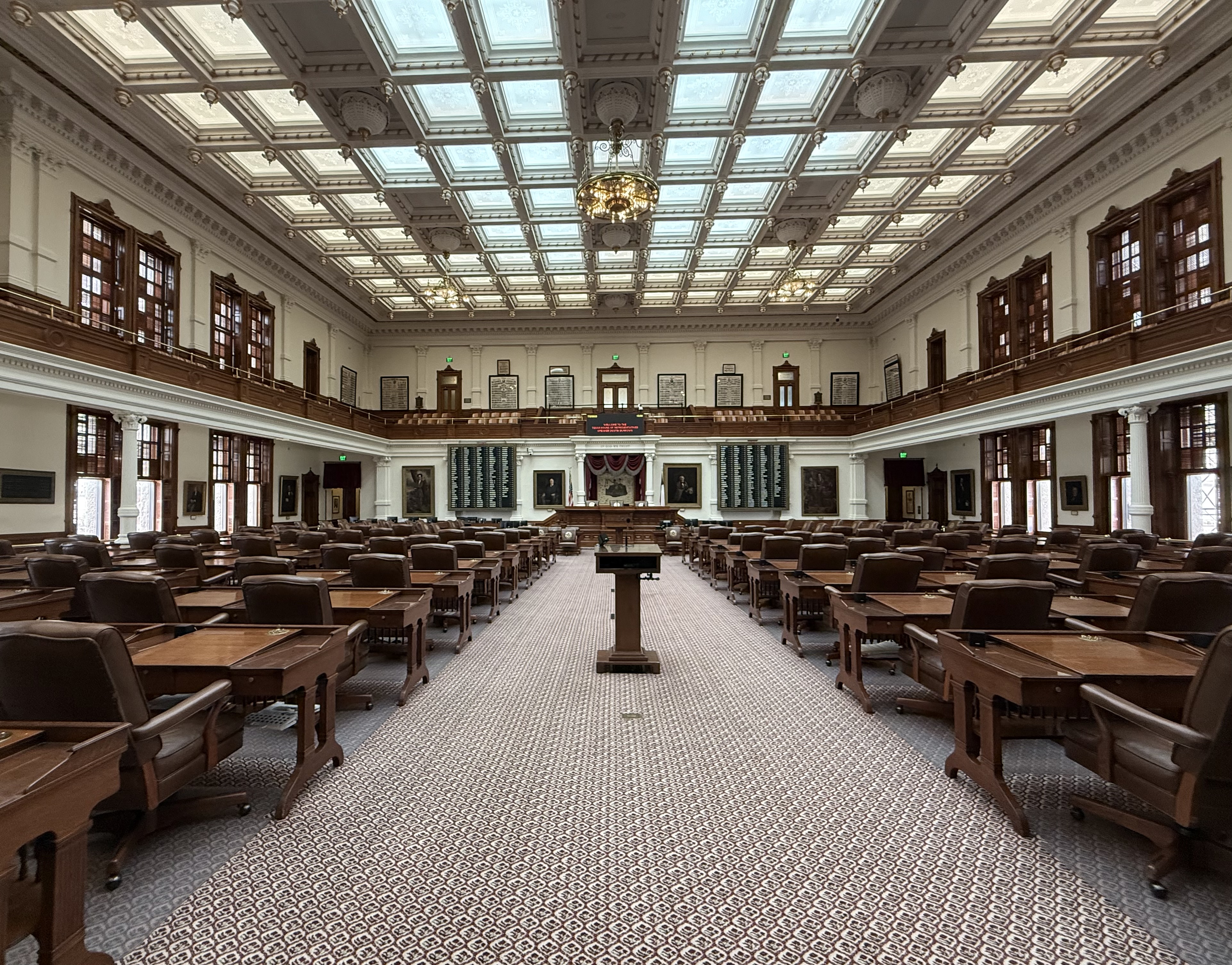 Empty legislative chamber with rows of wooden desks and a skylight ceiling.