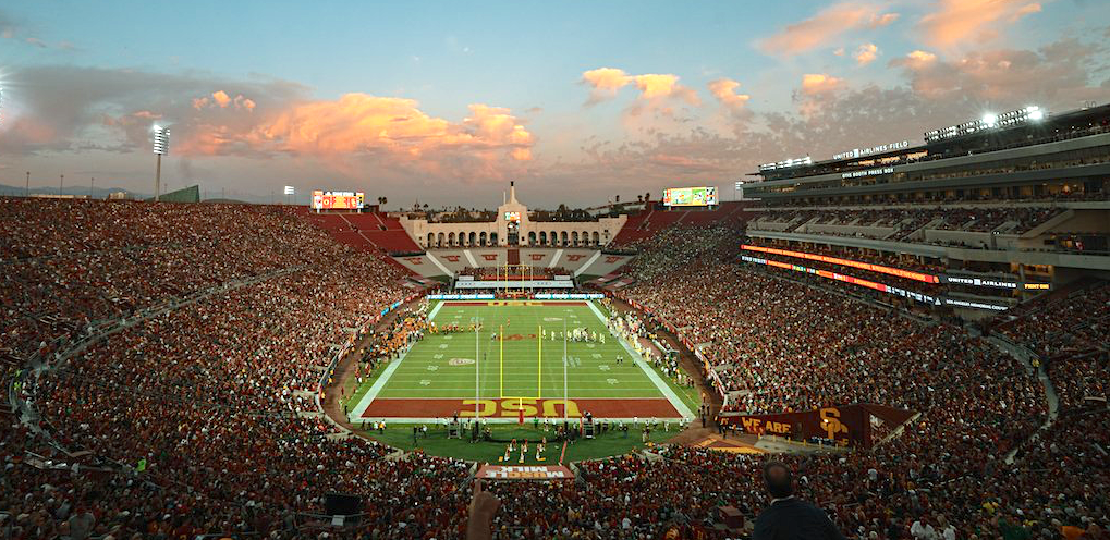 Wide-angle interior view of a packed football stadium at dusk with field lights on.