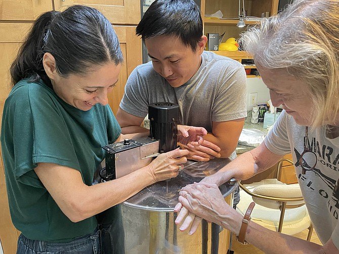 Three people pressing down on a metal honey extractor in a kitchen.