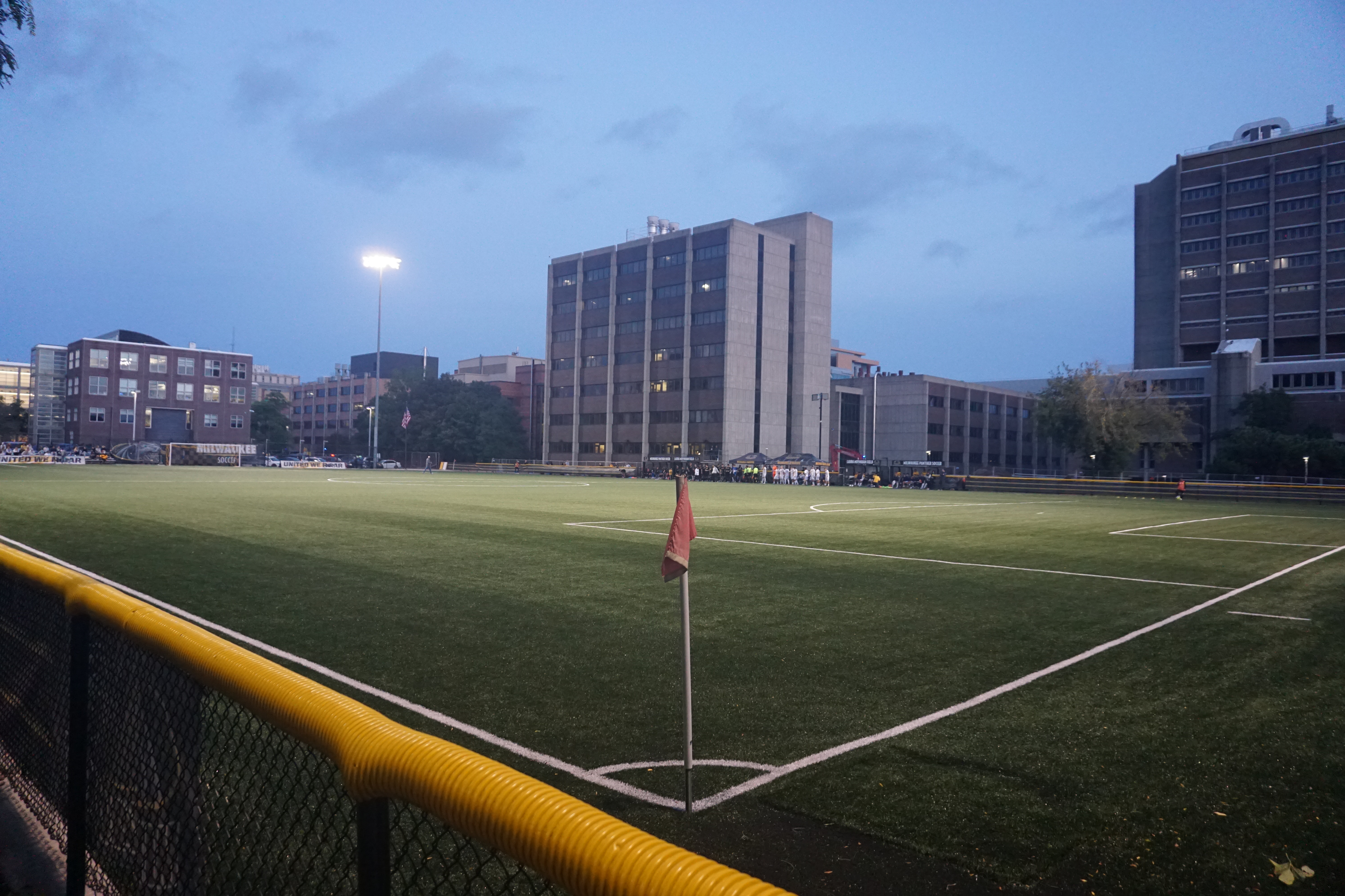Soccer field at dusk with a red corner flag, lit stadium lights, and urban buildings in the background.