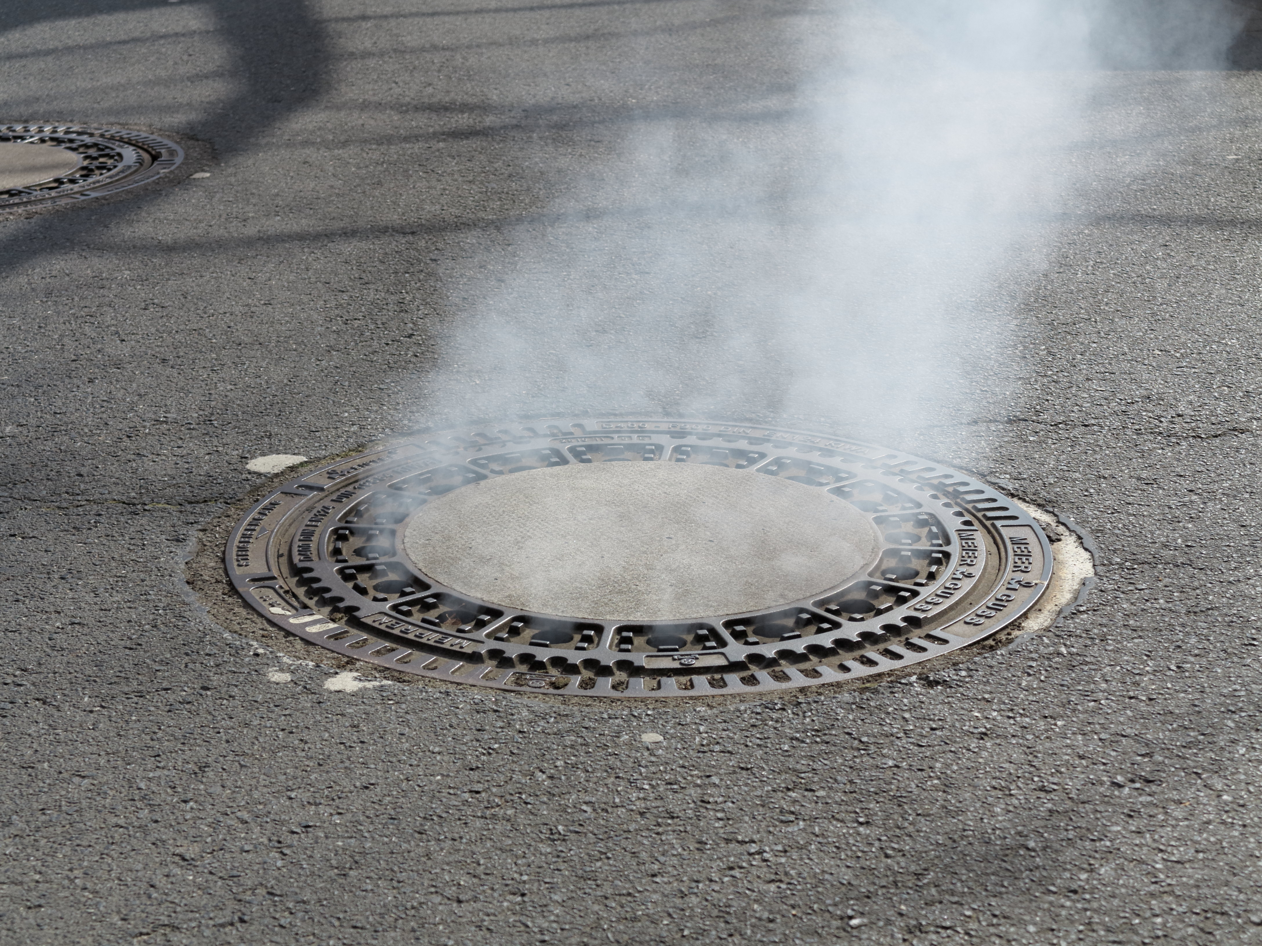 White smoke rises from a circular cast-iron manhole cover on an asphalt road.