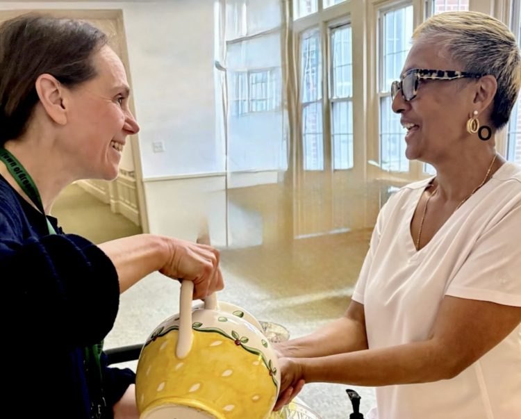 Two women smiling during a hand-blessing ceremony, one holding a decorative bowl indoors.