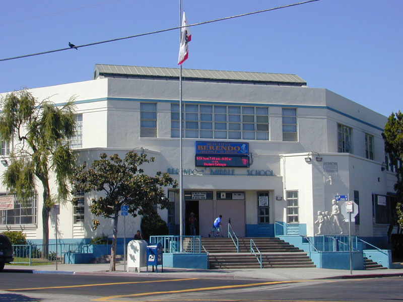 Exterior of a two-story white Art Deco school building with blue trim and a flagpole.