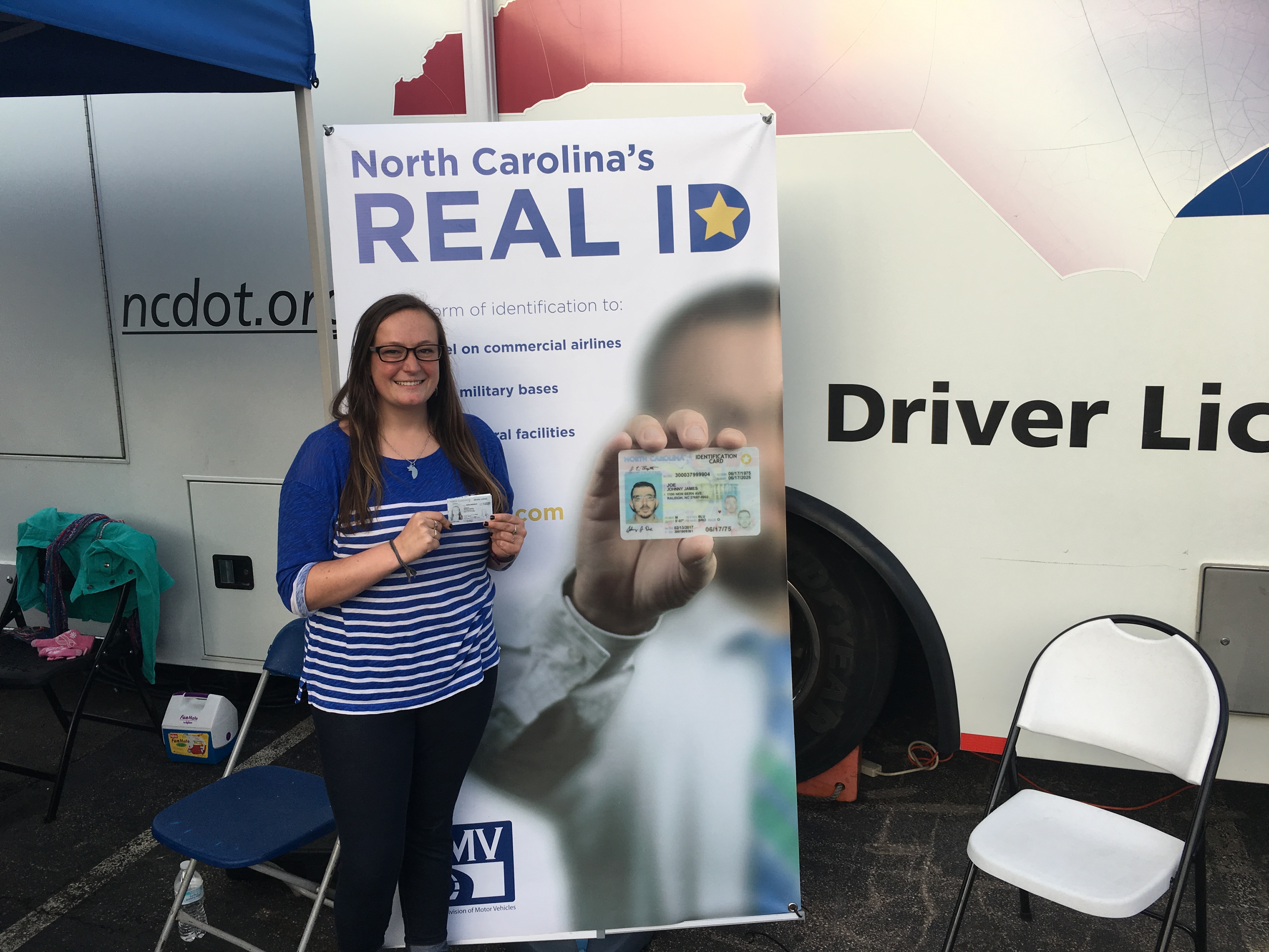 A woman holds up a driver's license in front of a DMV promotional banner.