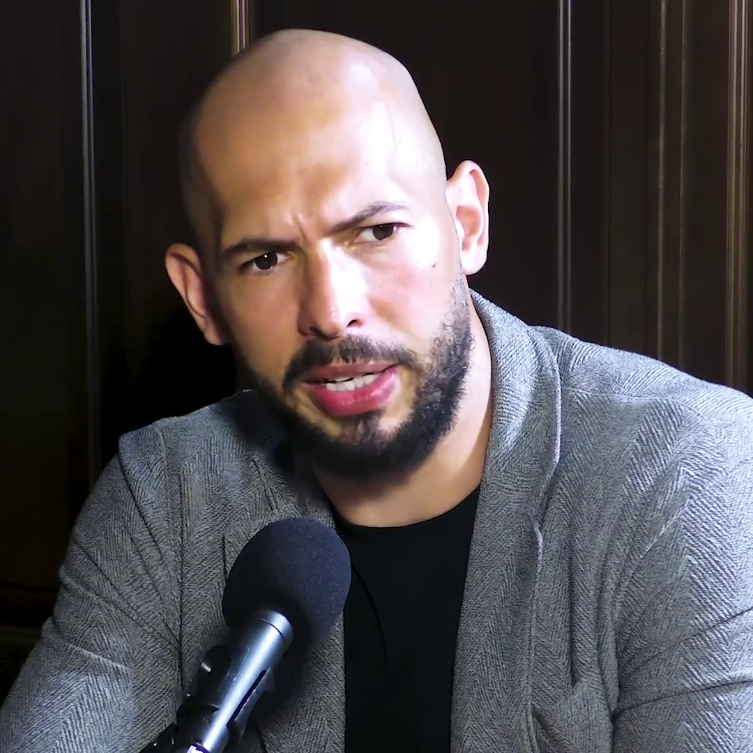 Bald man with short beard in grey blazer seated at a podcast microphone.