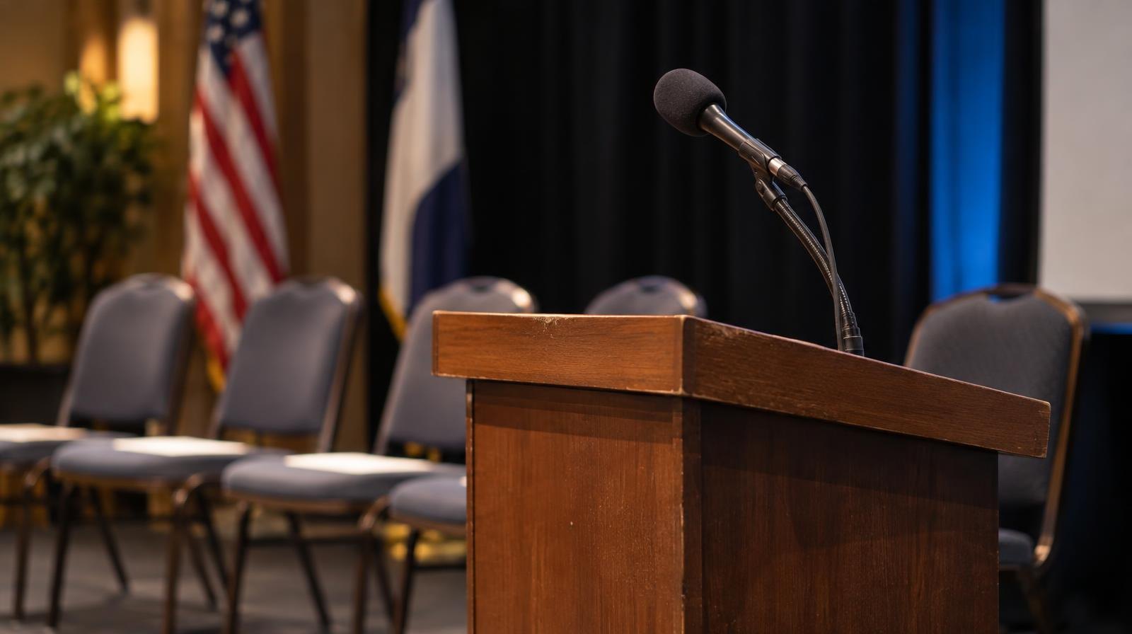 A wooden podium with a microphone in a softly lit auditorium with empty chairs.