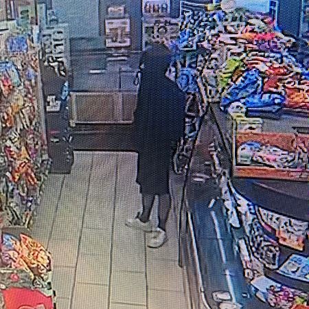 Low-resolution security camera still of a person in a black hoodie standing in a convenience store aisle.