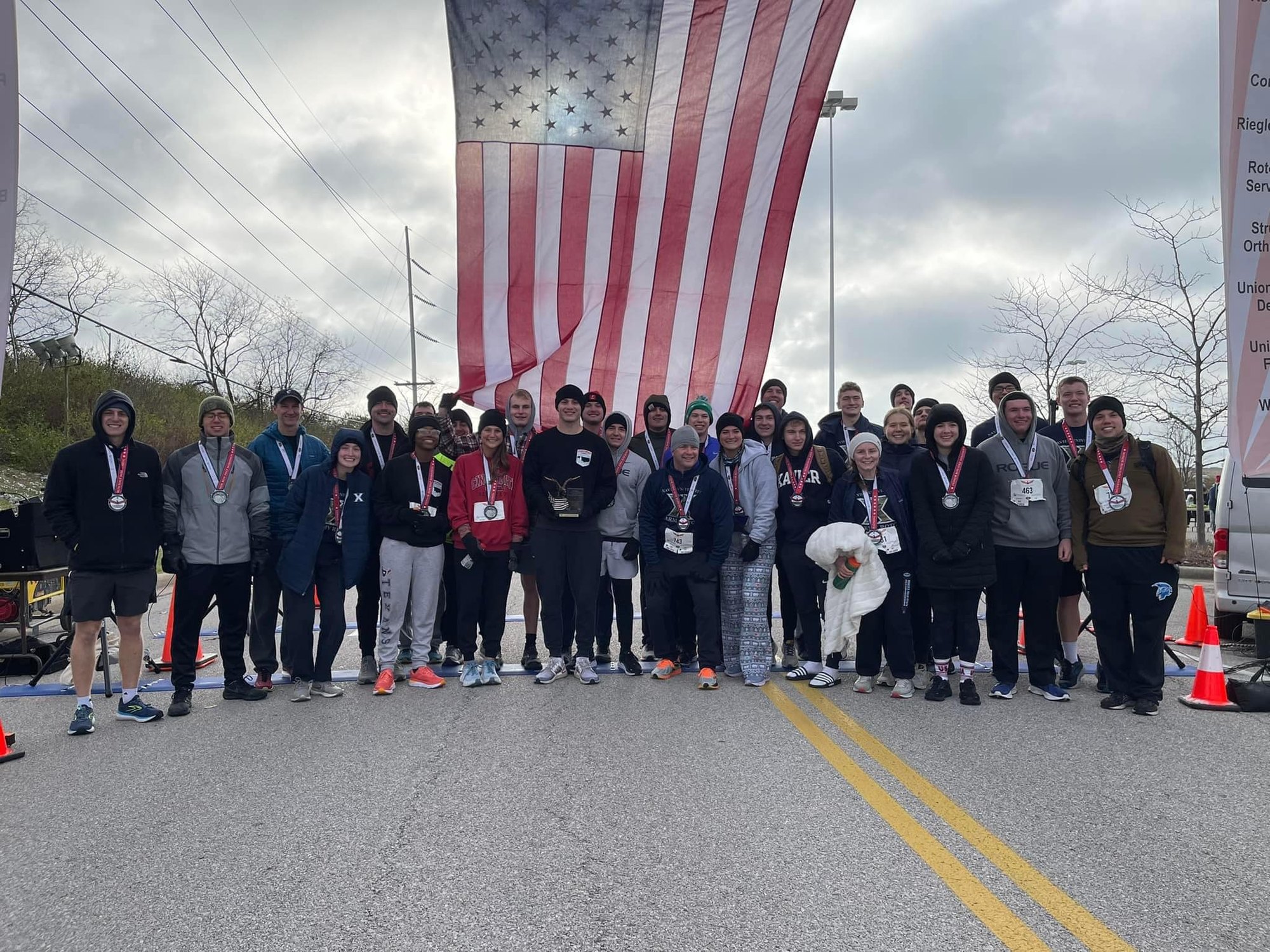 A group of race finishers wearing medals pose under a large American flag on a road.
