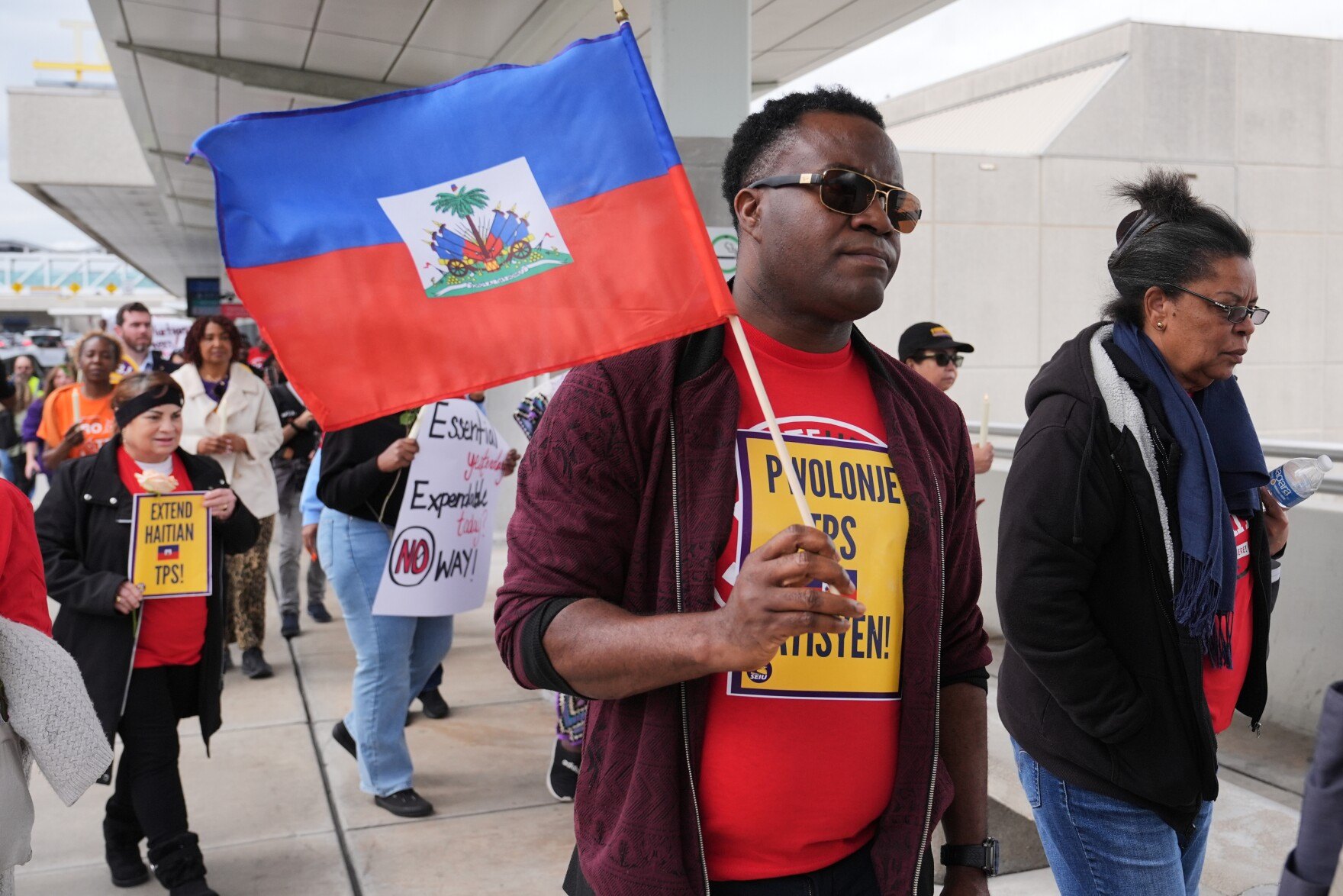 Man in sunglasses holds a Haitian flag at an outdoor rally with protesters holding TPS signs.