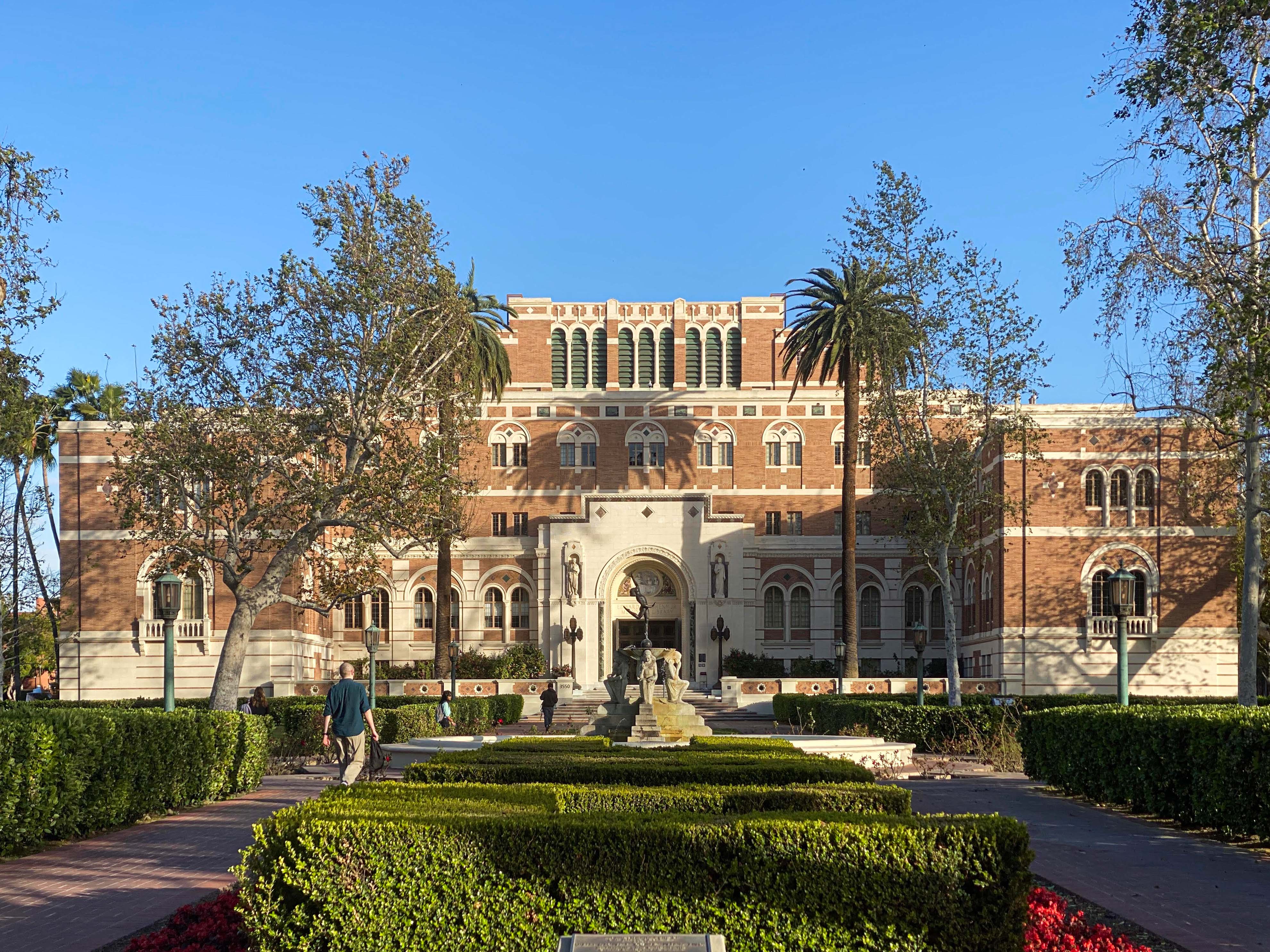 Red-brick Romanesque Revival library building on a sunny campus with palm trees and a central walkway.