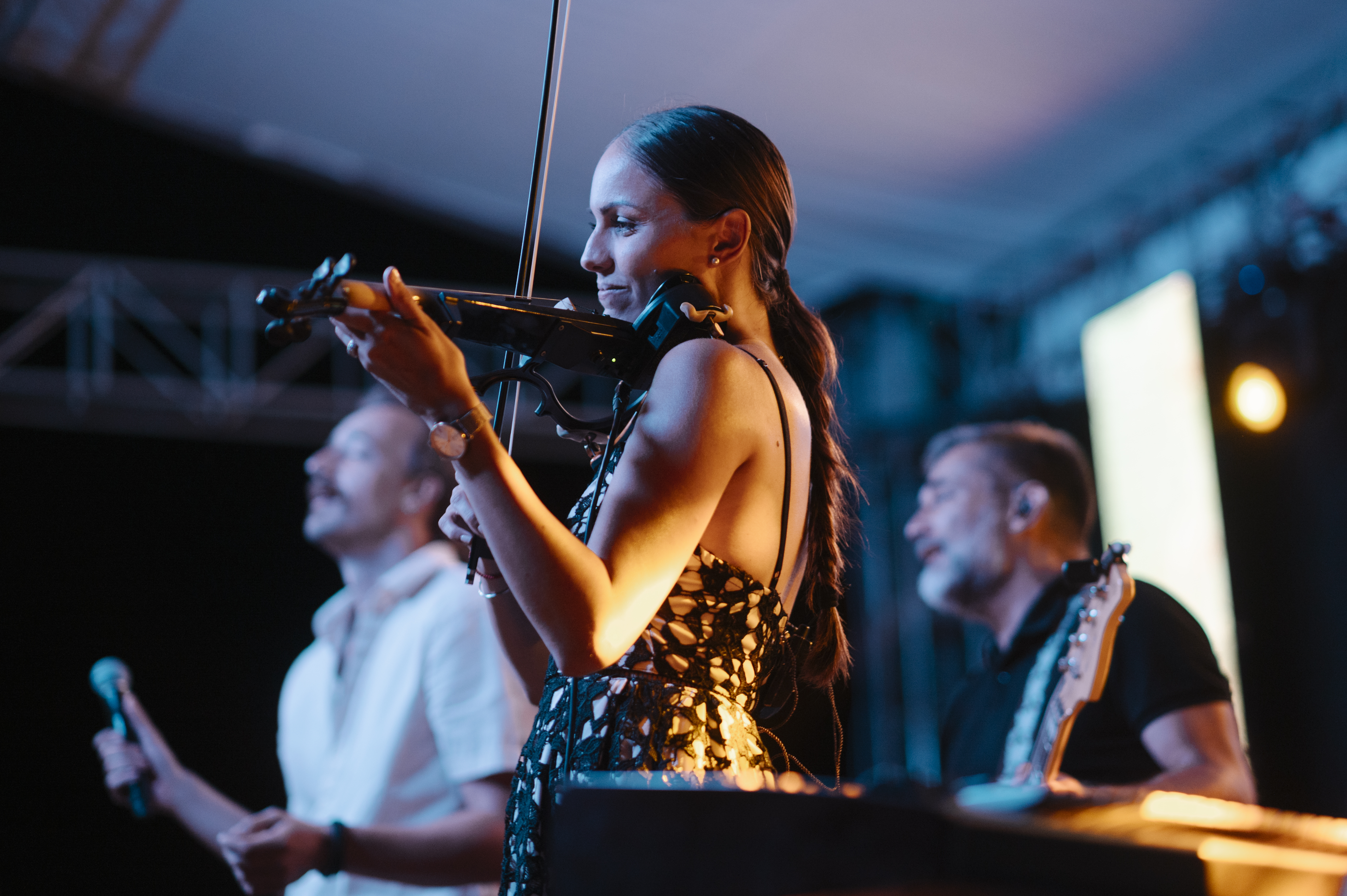 Musicians perform on a colorfully lit outdoor stage at dusk.