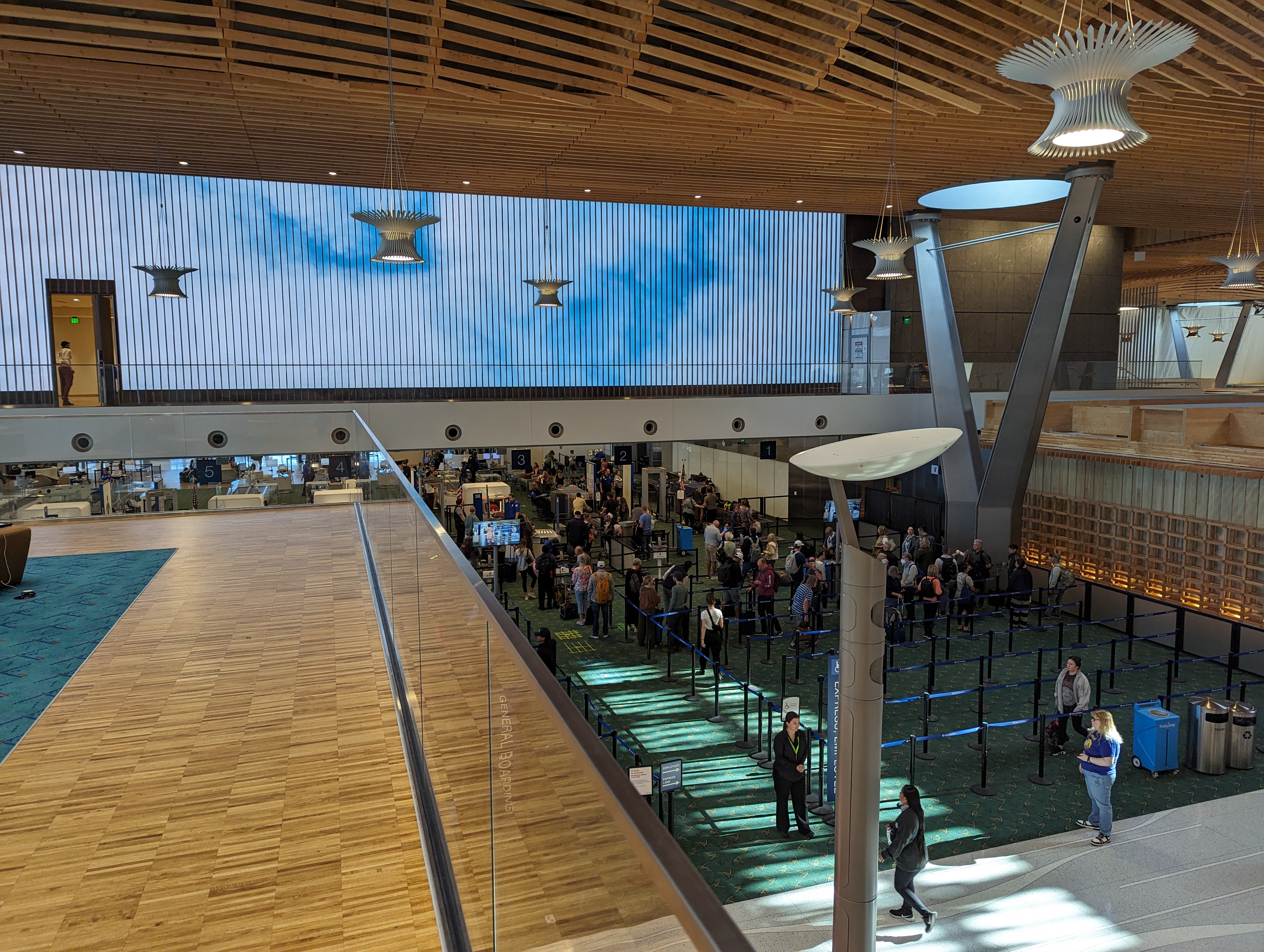 Airport security checkpoint viewed from above, with passengers queued beneath a large digital sky display.