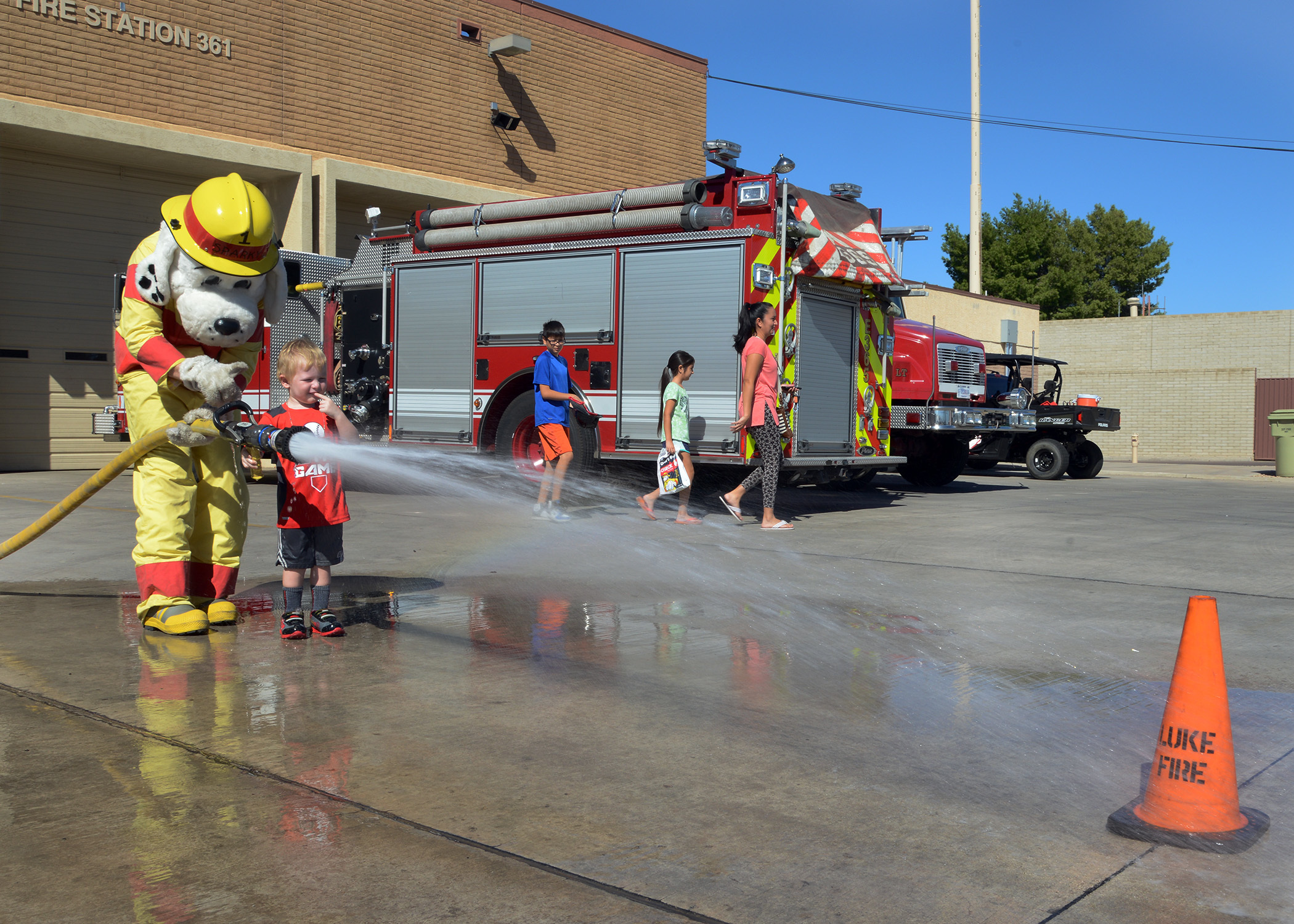 A child aims a fire hose with a costumed mascot at an outdoor fire station open house.