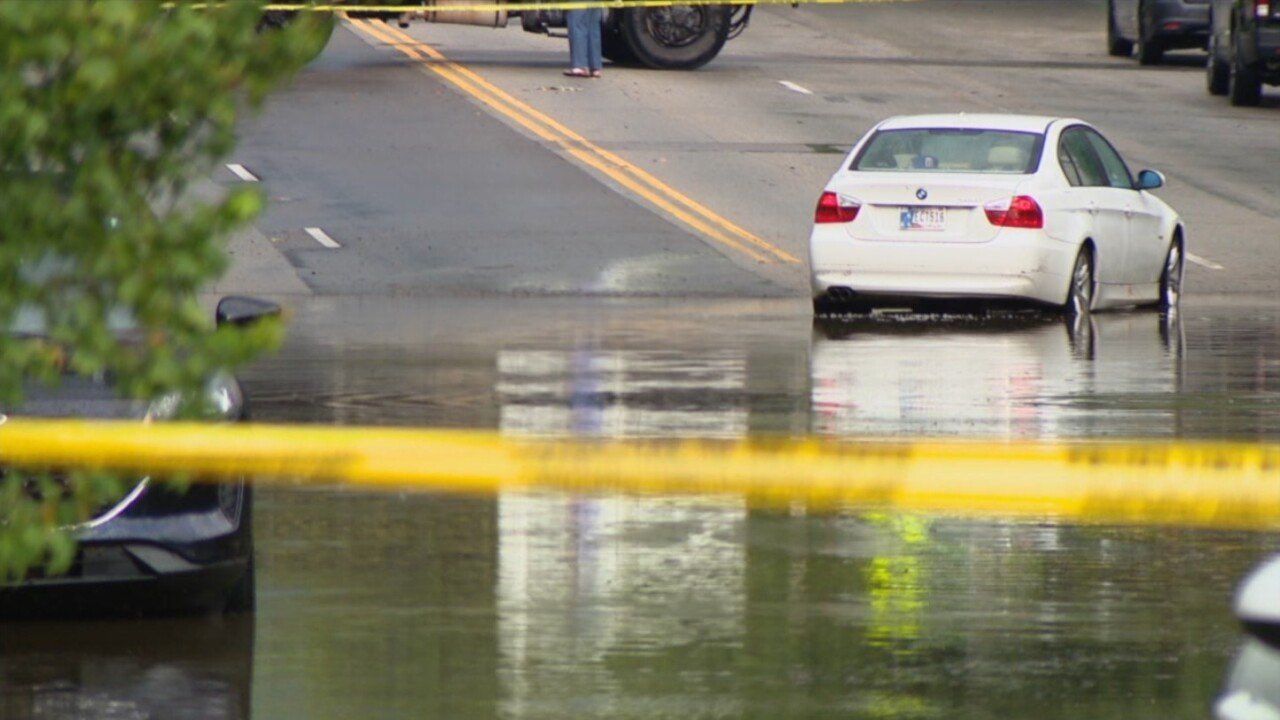 White sedan partially submerged in floodwater on a city street with caution tape in foreground.