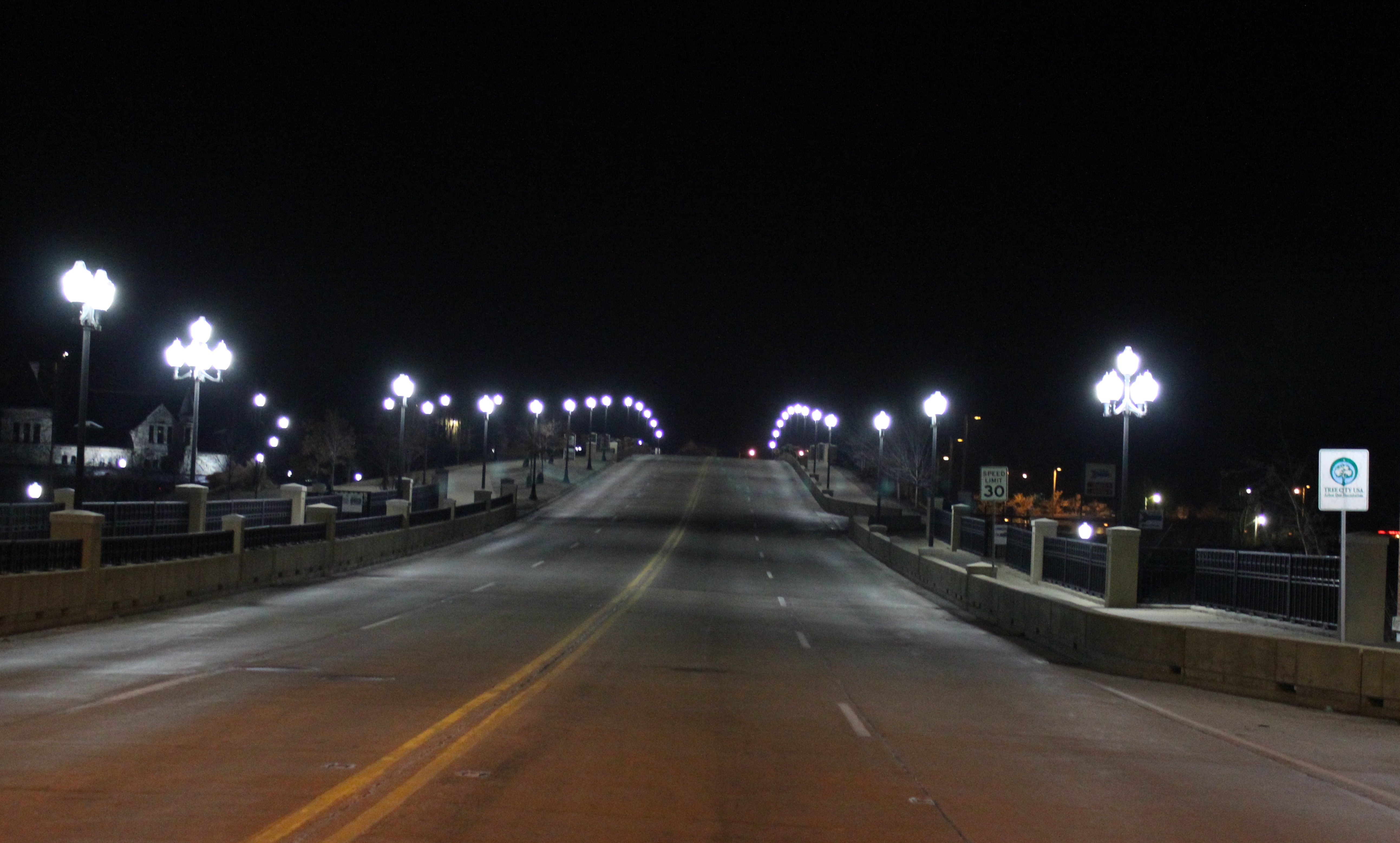 A wide bridge roadway at night, lined with ornamental street lamps on both sides.