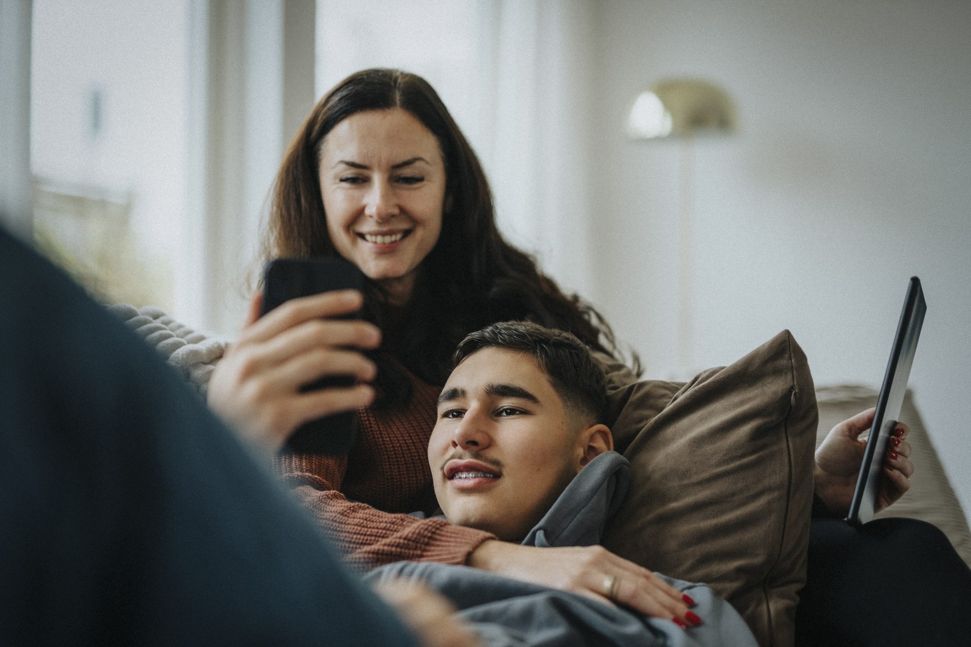 Teenage boy sharing smart phone with mother while lying on sofa in living room at home