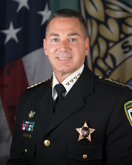 Law enforcement official in dark dress uniform with gold sheriff's star badge and service ribbons.