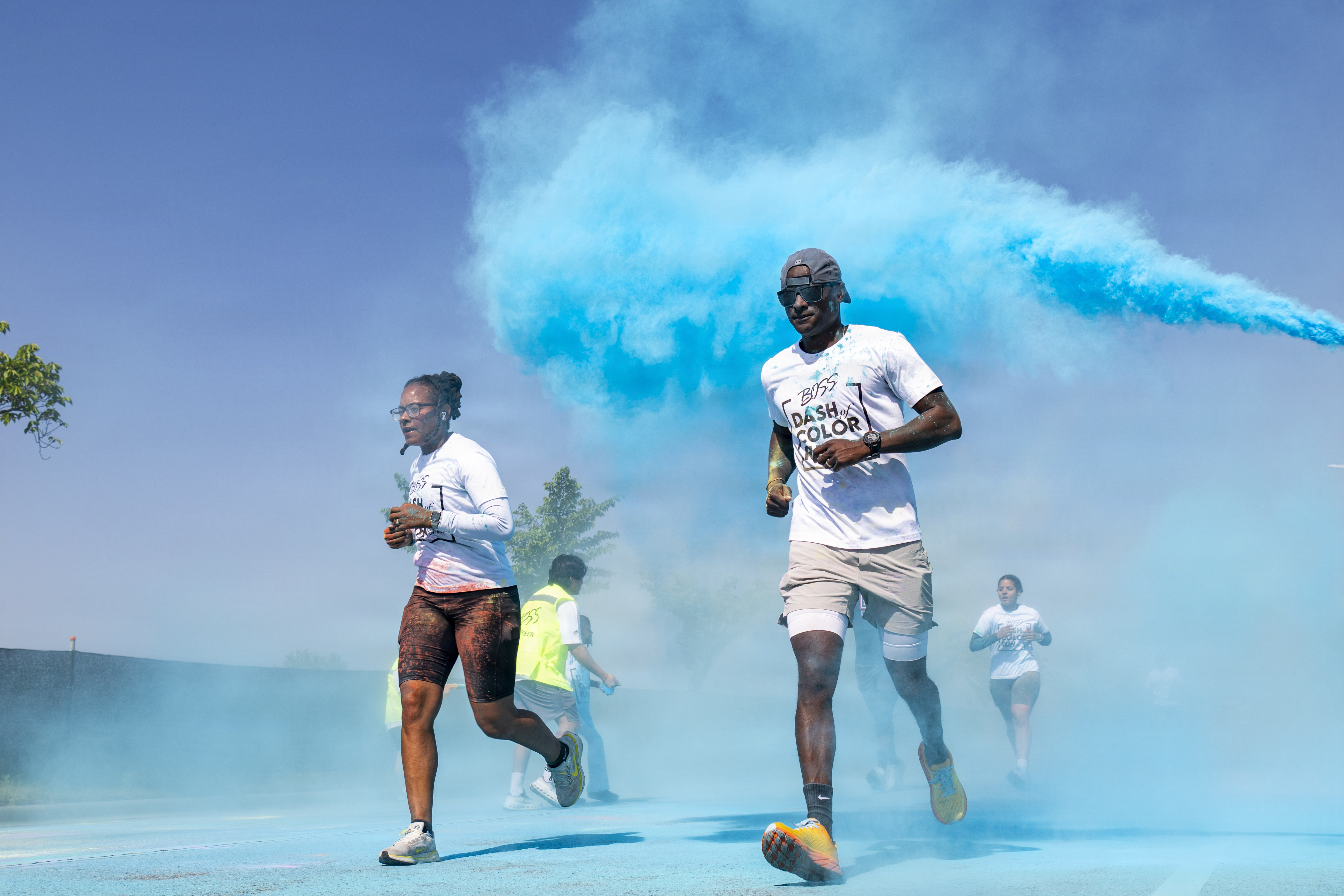 Runners in white shirts pass through a large blue powder cloud outdoors.