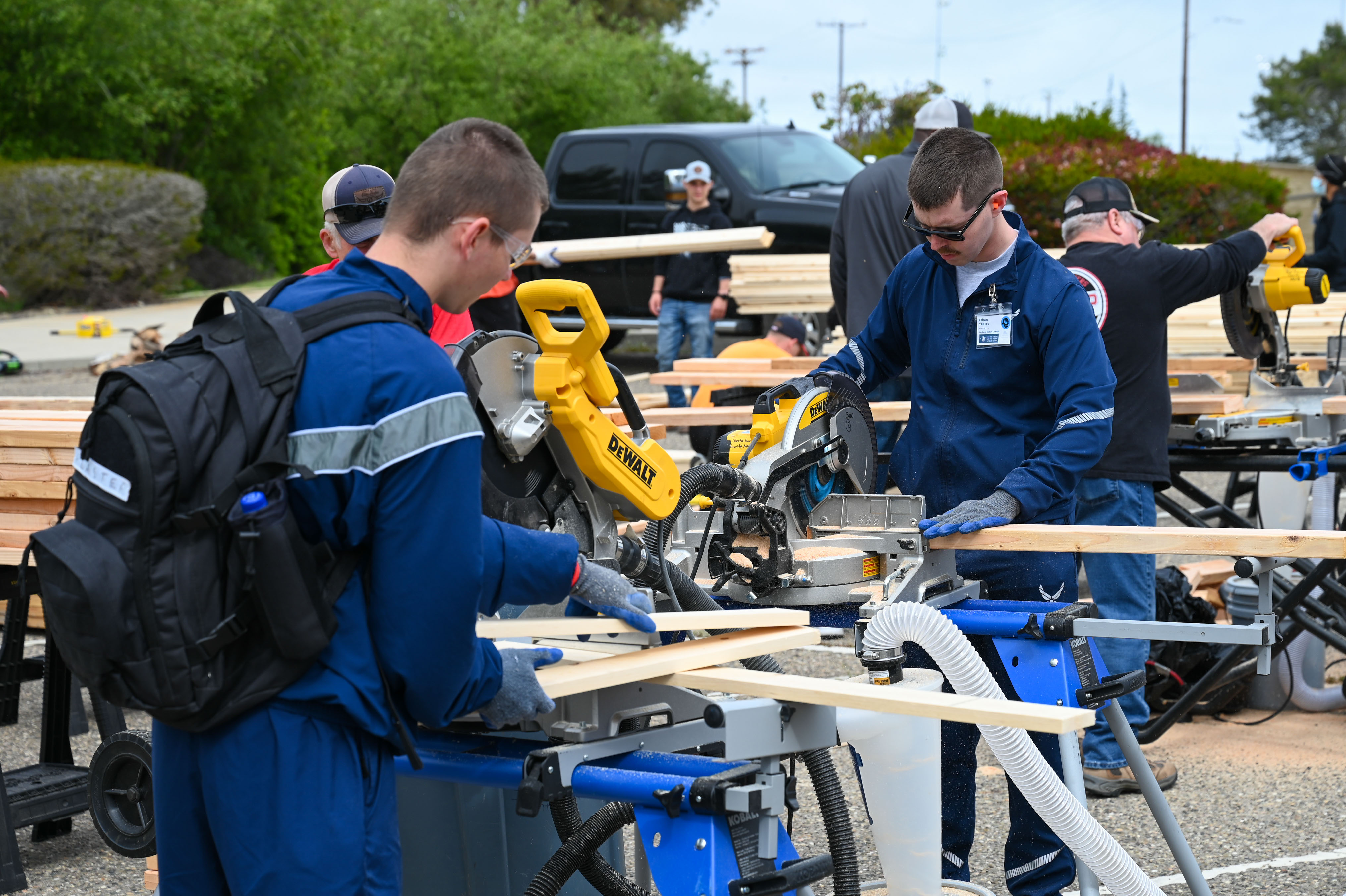 Volunteers in blue uniforms cut lumber with a power saw at an outdoor bed-building event.