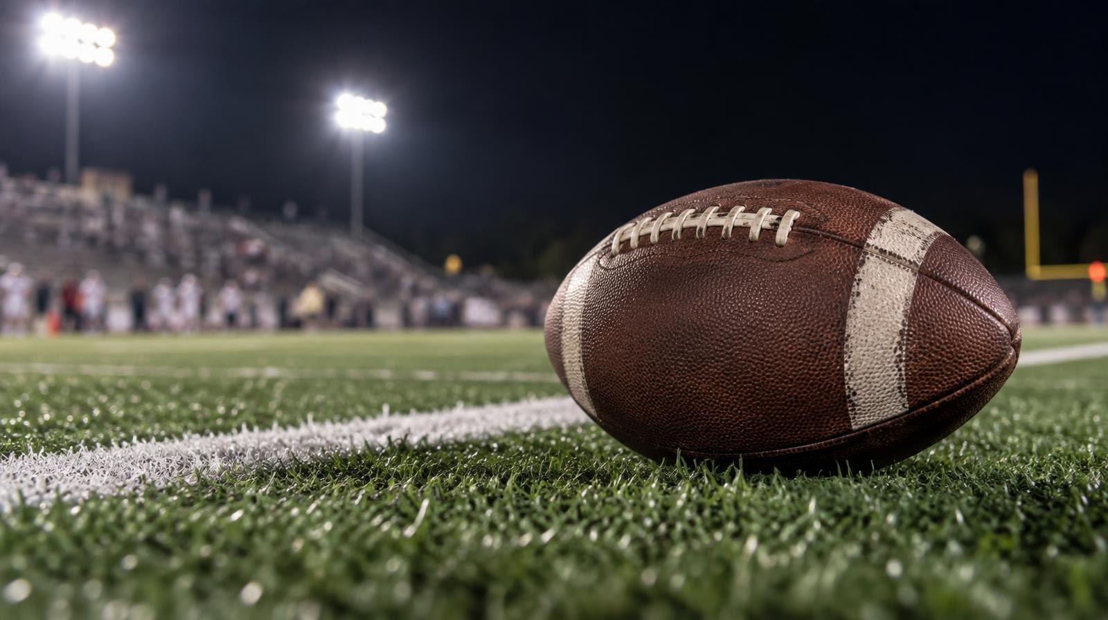 An American football on green turf beside a white yard line under bright lights.