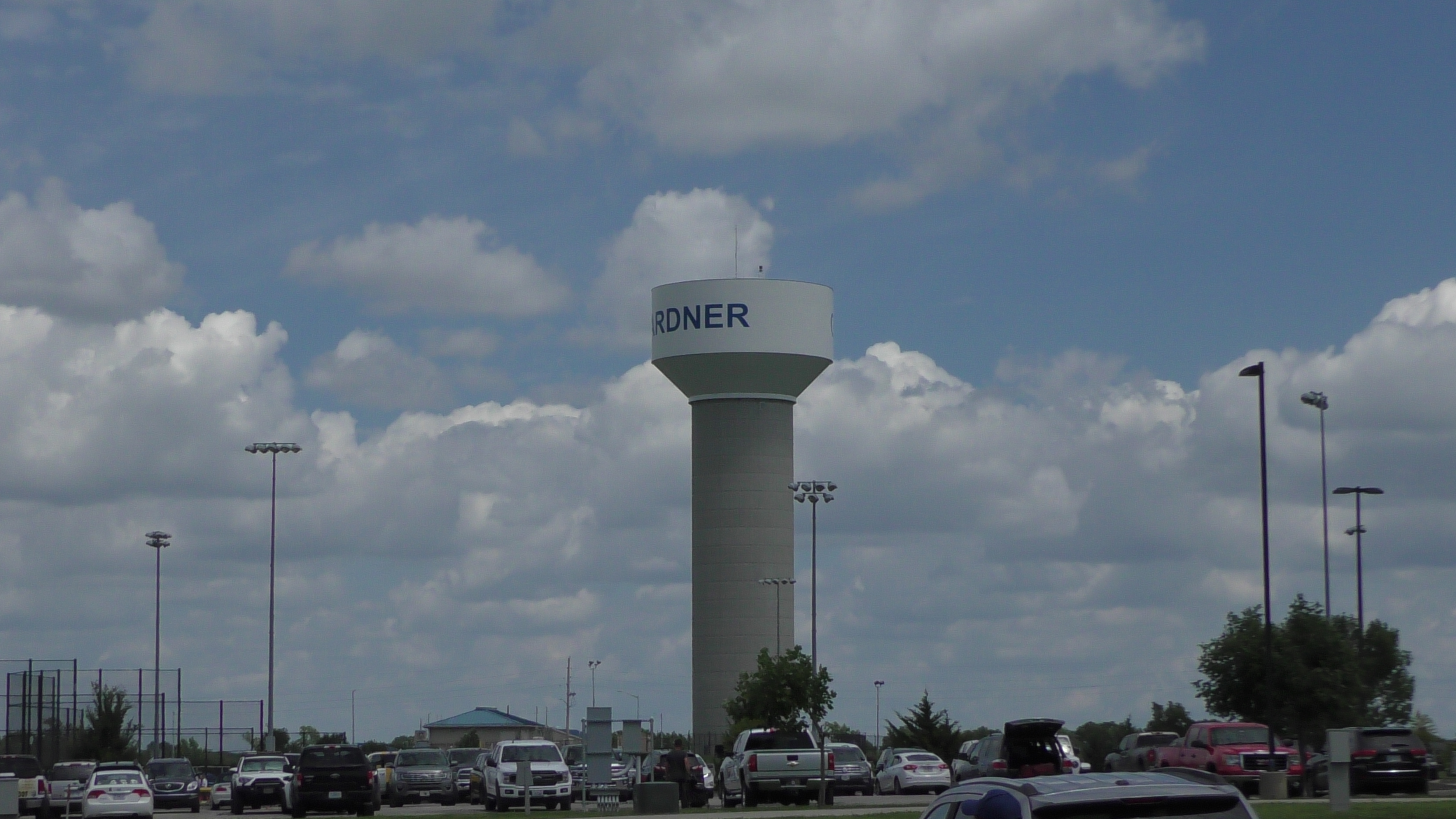 Tall cylindrical water tower with city name visible, surrounded by a parking lot and light poles.