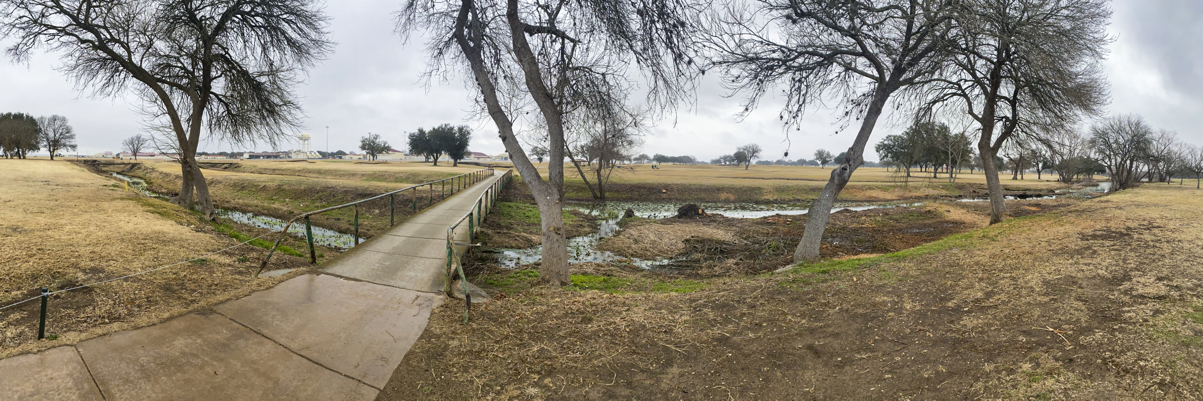 Concrete path leads to a small footbridge over a drainage channel beside a detention pond.