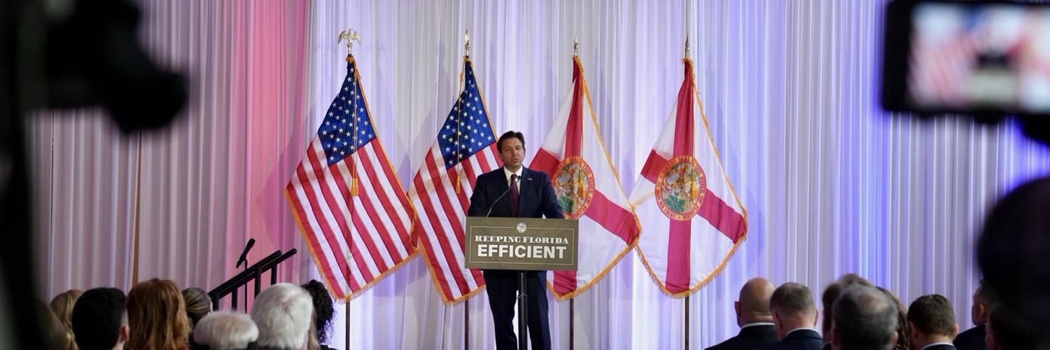 Man in suit stands at a podium flanked by American and Florida state flags.