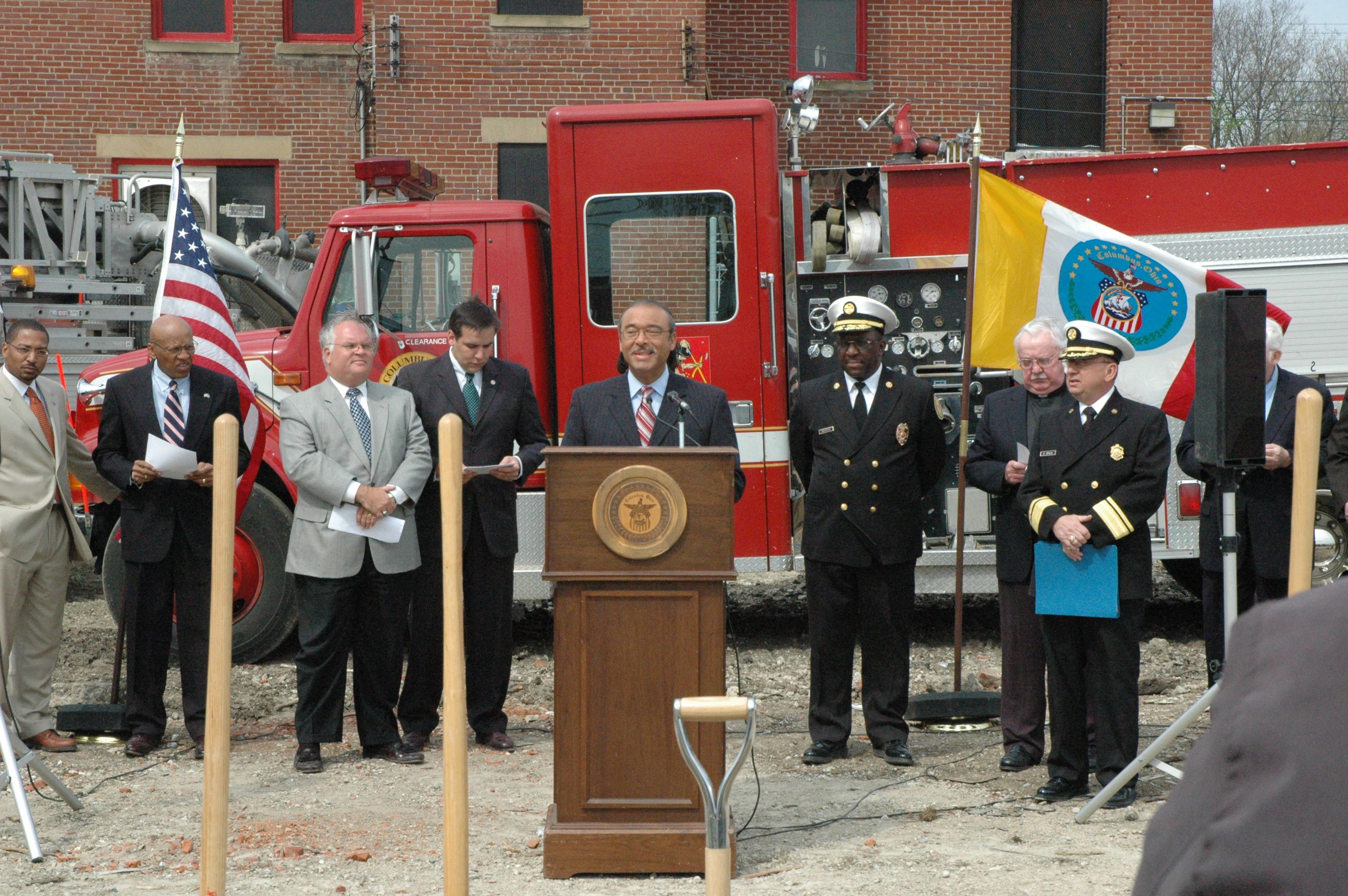 A man speaks at a lectern at an outdoor fire station groundbreaking ceremony with firefighters and fire trucks behind him.