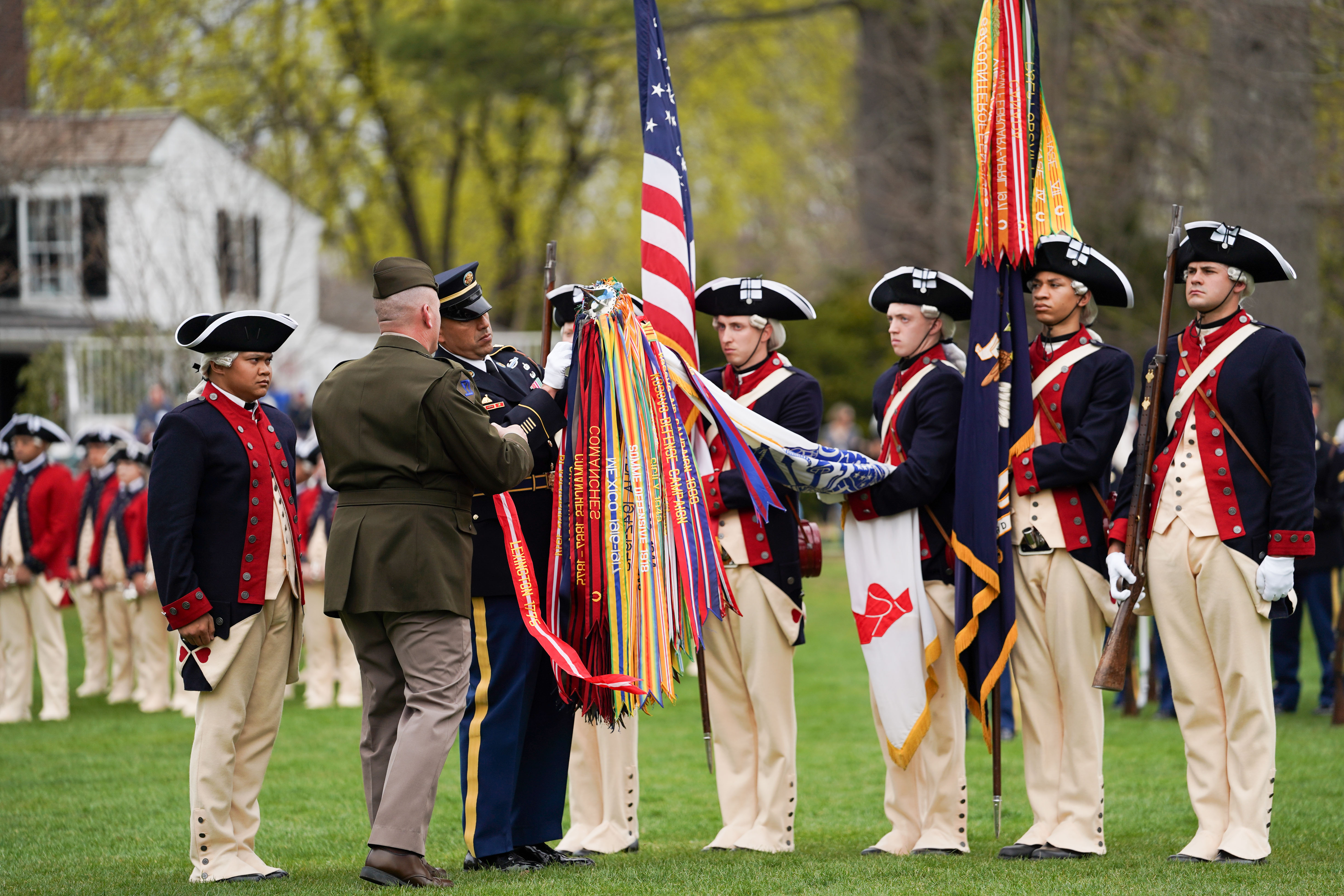 Soldiers in Continental Army uniforms stand in formation on a green lawn with historic buildings behind them.