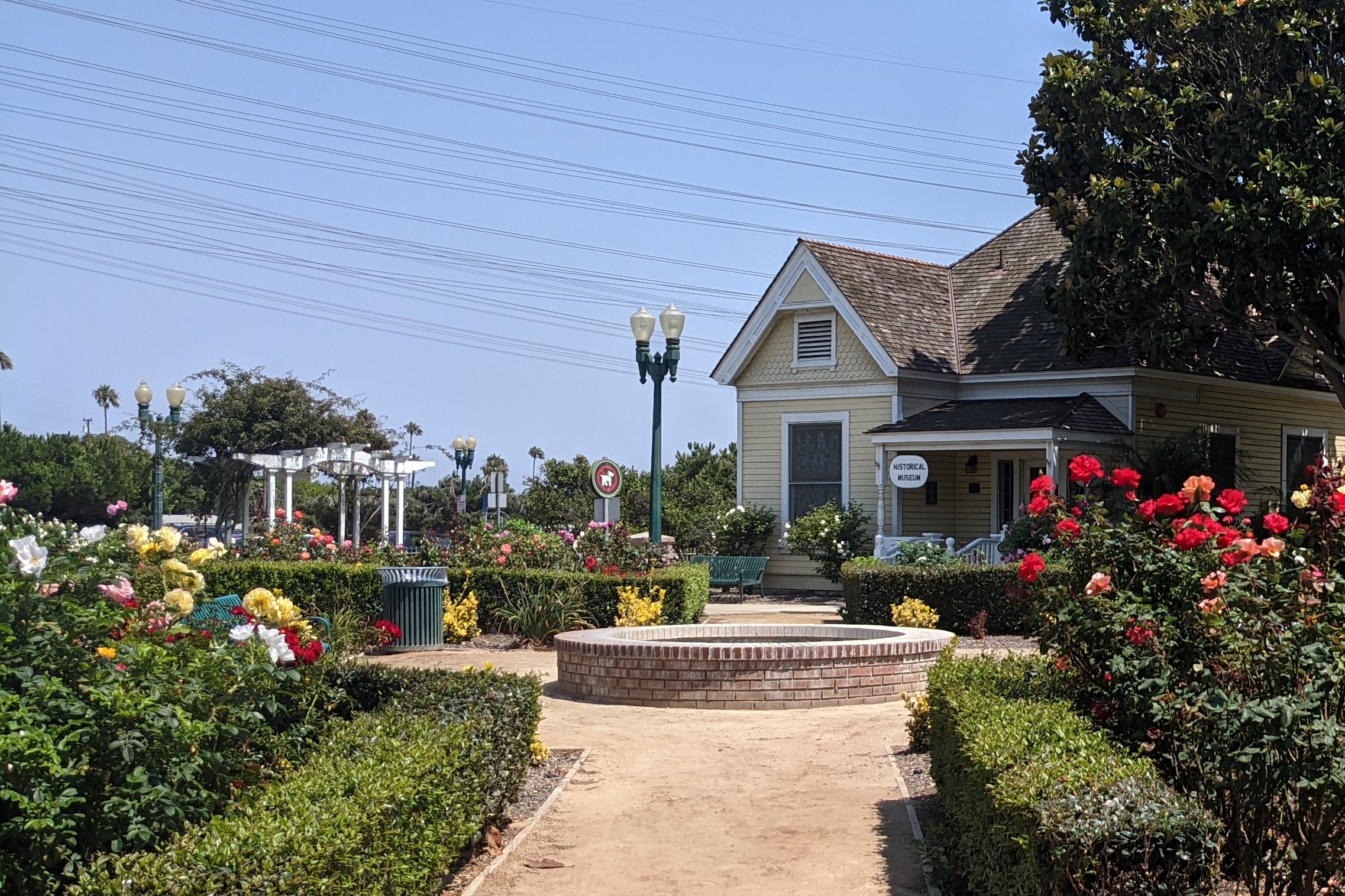 A landscaped public park with a dry fountain, rose garden path, and small yellow cottage building.