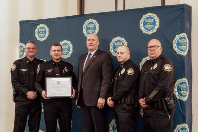 Five men in police uniforms and a suit pose holding a framed certificate in front of a law enforcement backdrop.