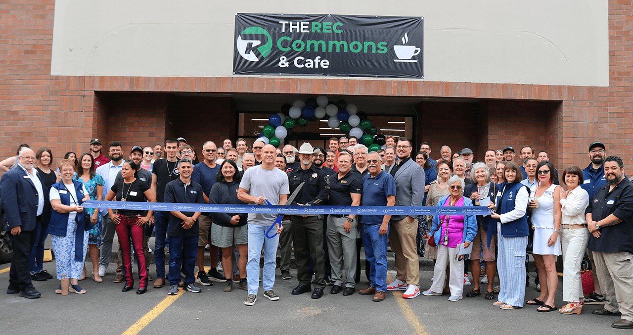 Crowd gathered outside a brick building for a ribbon-cutting ceremony with blue ribbon and balloons.