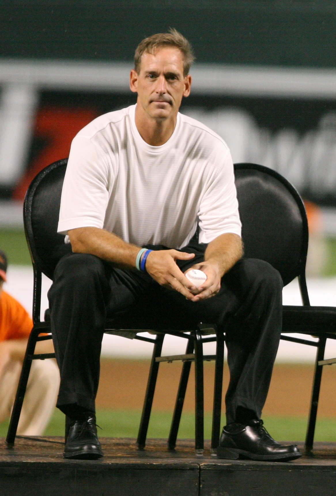 Man in white t-shirt seated on a baseball field holding a baseball.