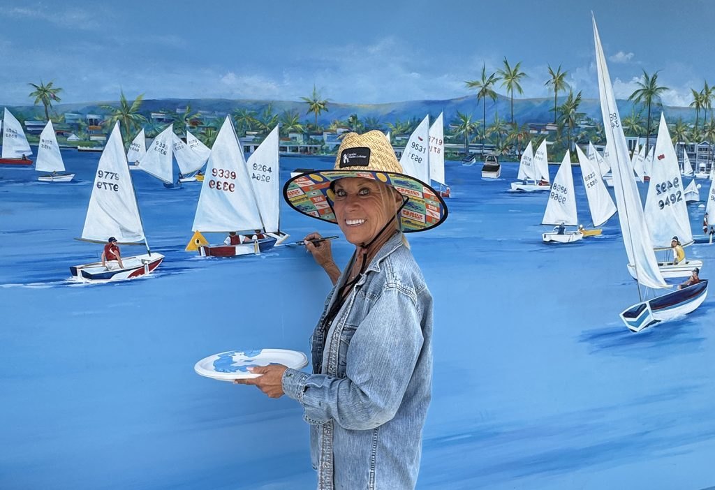 Woman in straw hat holding paint palette and brush in front of a large sailboat mural.