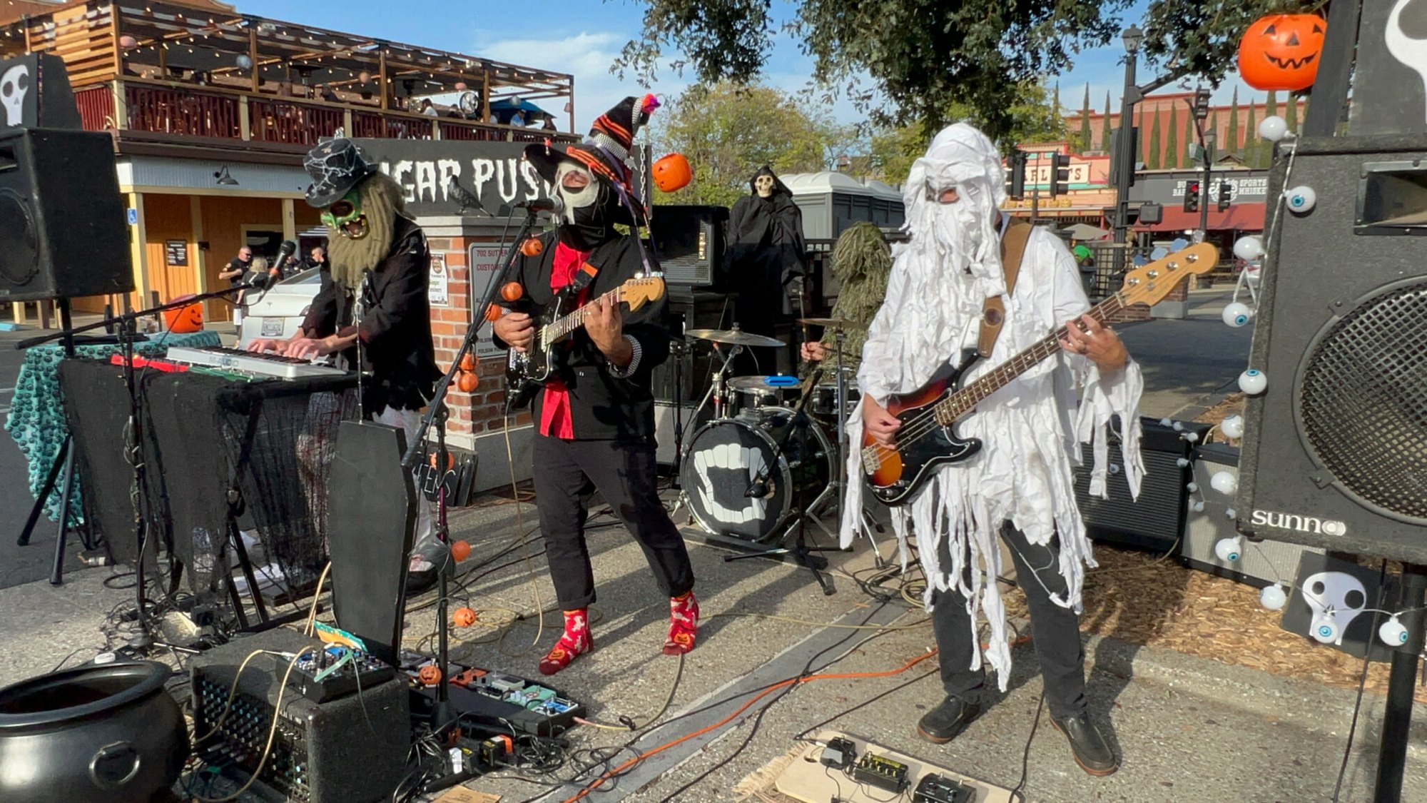 Four musicians in Halloween costumes perform outdoors with guitars, keyboard, and drums.
