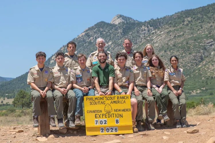 Group of scouts and adults in tan uniforms posing before a yellow Philmont Scout Ranch sign with mountains behind them.