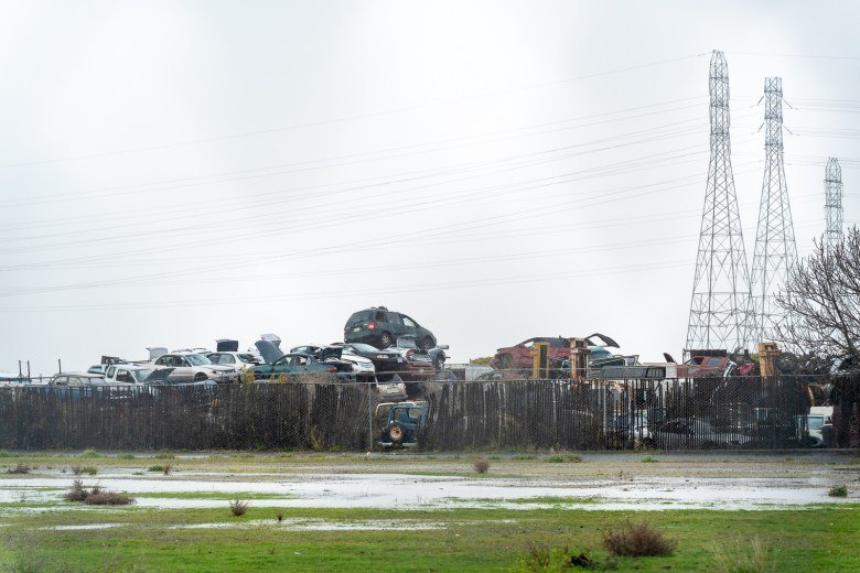 Stacked vehicles visible over a fence at a salvage yard with transmission towers and overcast sky.