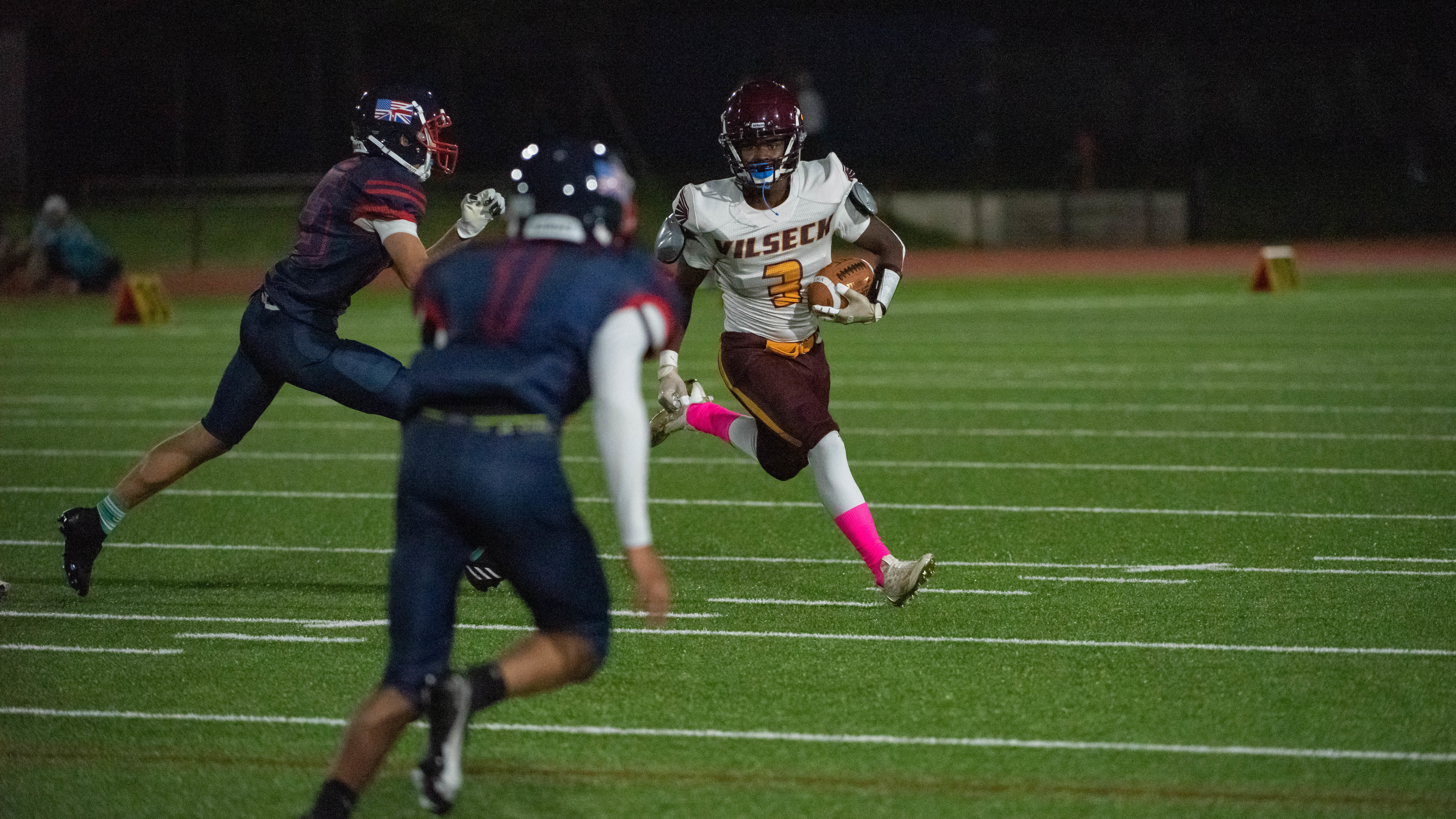 High school football players tackling during a night game on artificial turf under stadium lights.
