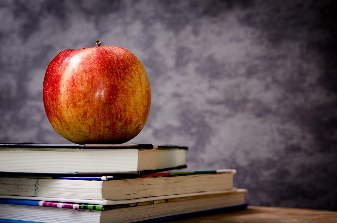 A red and yellow apple rests on a stack of hardcover books against a gray background.