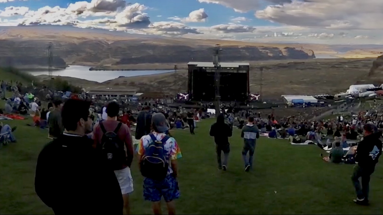 Festival crowd on a grassy slope facing an outdoor stage with a river gorge behind.
